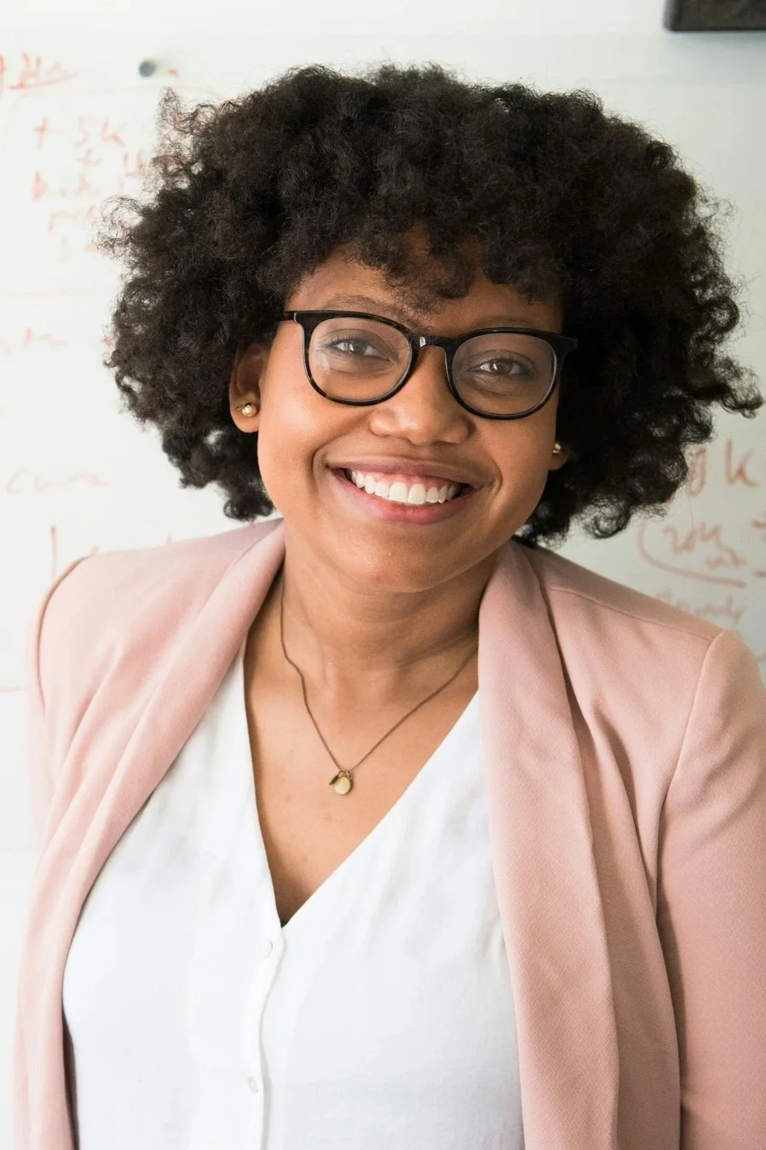 Close-up of a smiling woman with curly black hair, wearing glasses, a light pink blazer, a white blouse, and a necklace, with a whiteboard in the background.