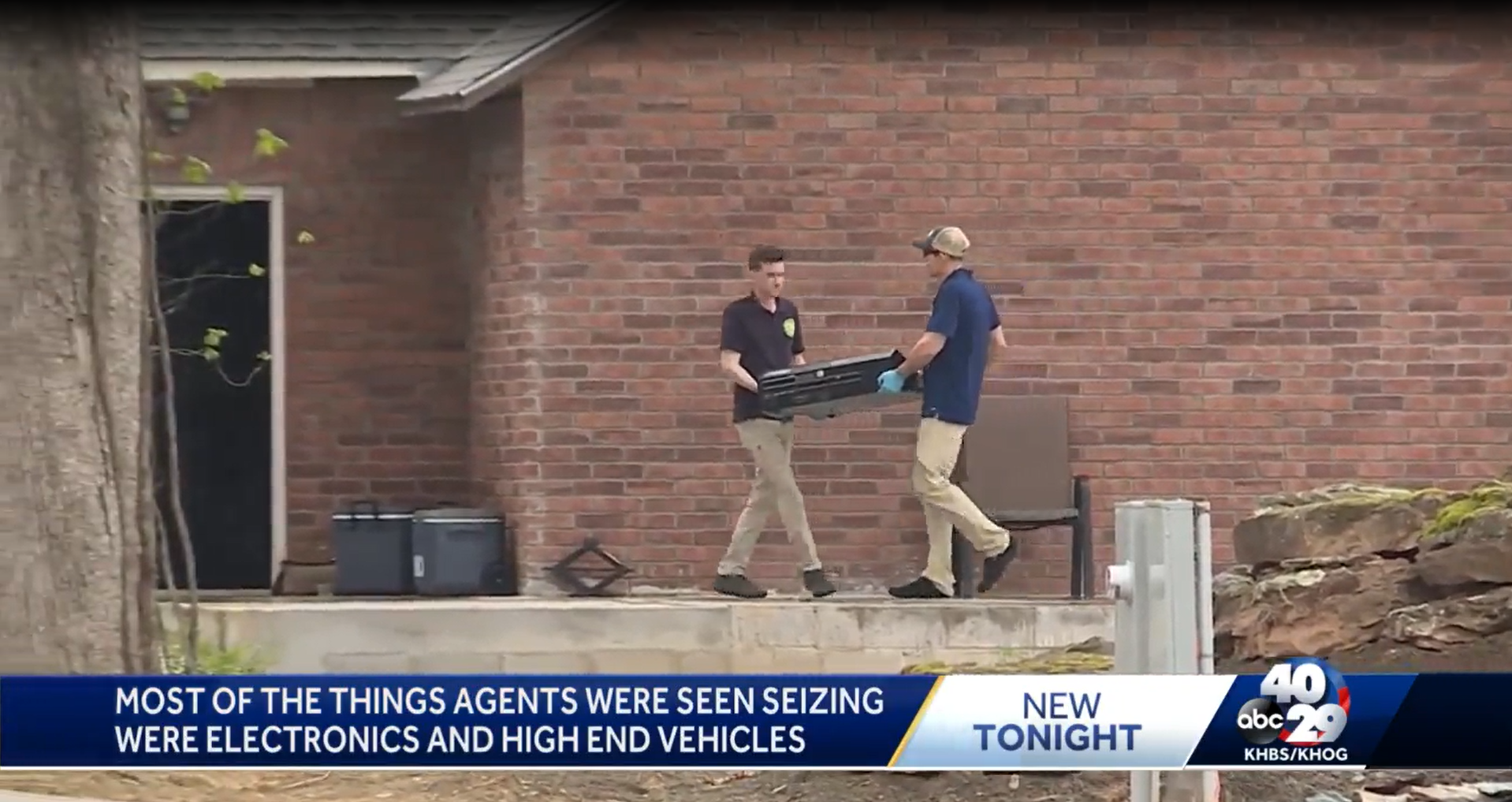 Two agents are seen seizing electronics outside a brick building. One agent wearing a black shirt and tan pants, the other wearing a blue shirt, tan pants, and a cap. They are handling a black electronic device. There is a bench, some equipment, and a rock garden nearby. A news broadcast overlay at the bottom states, "Most of the things agents were seen seizing were electronics and high end vehicles..."