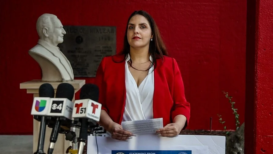 A woman in a red blazer holding papers stands behind a podium with multiple microphones, with a red wall and a statue of a man in the background.