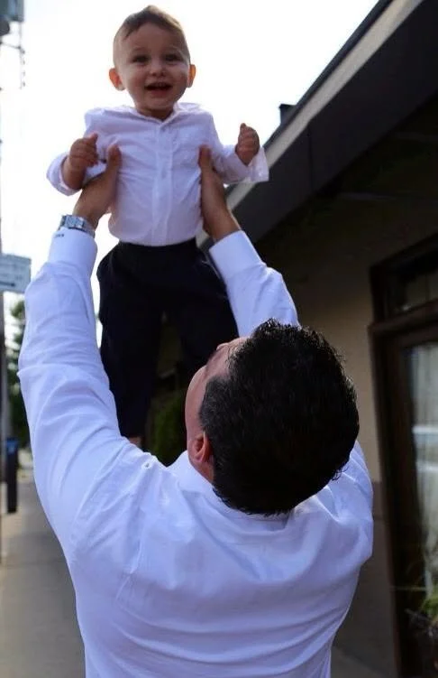 Michael Castillero lifting his infant son overhead, photographed from behind, 2013