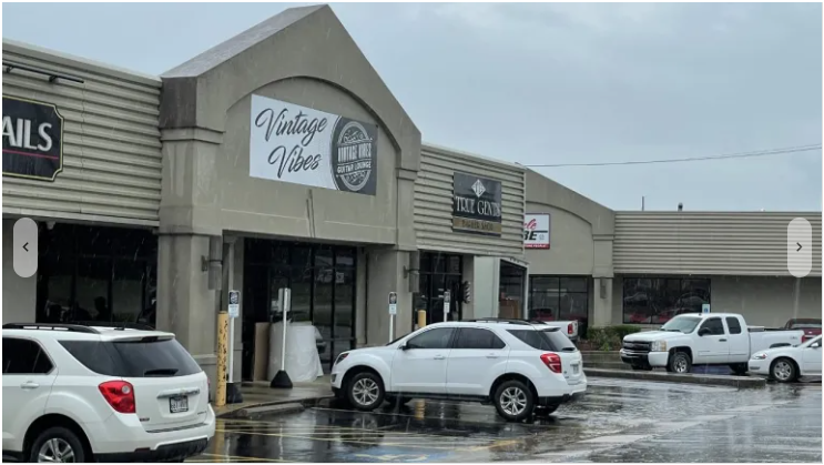 A shopping plaza with several storefronts, including one named Vintage Vise. There are multiple cars parked in the wet parking lot on a rainy day.