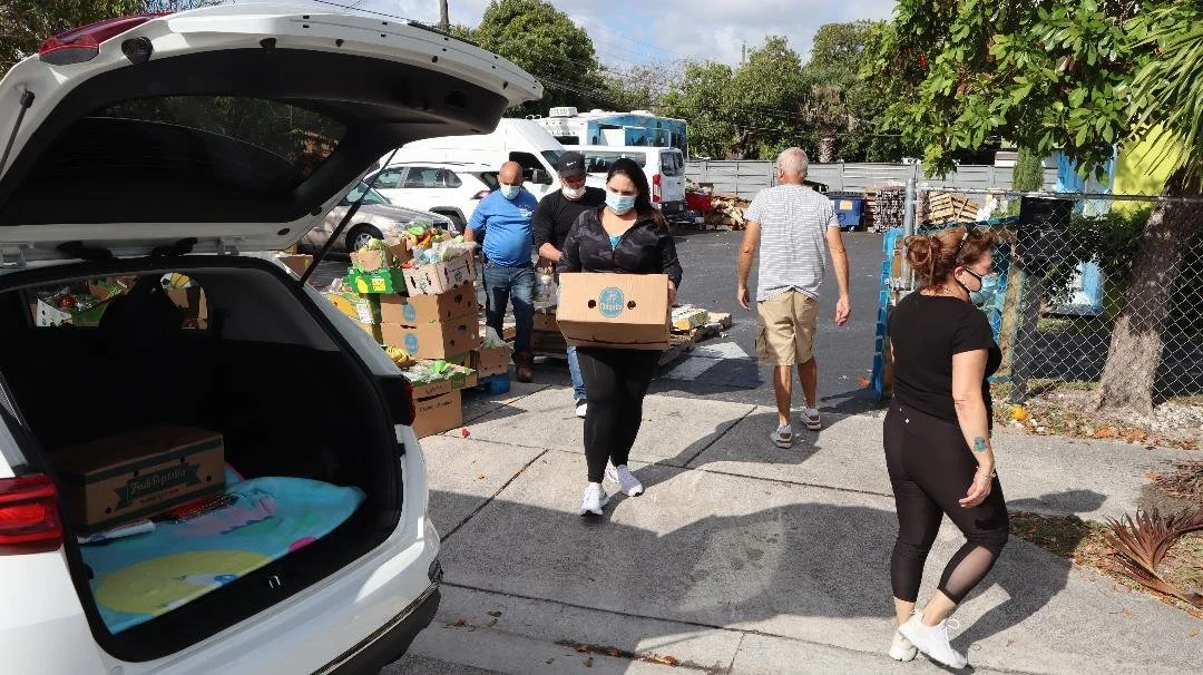 People wearing masks unload boxes of food and supplies from a car at an outdoor distribution event.