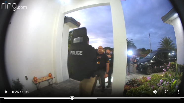 Police officers and a person in police gear outside a house, with a black vehicle and palm trees in the background, during dusk.