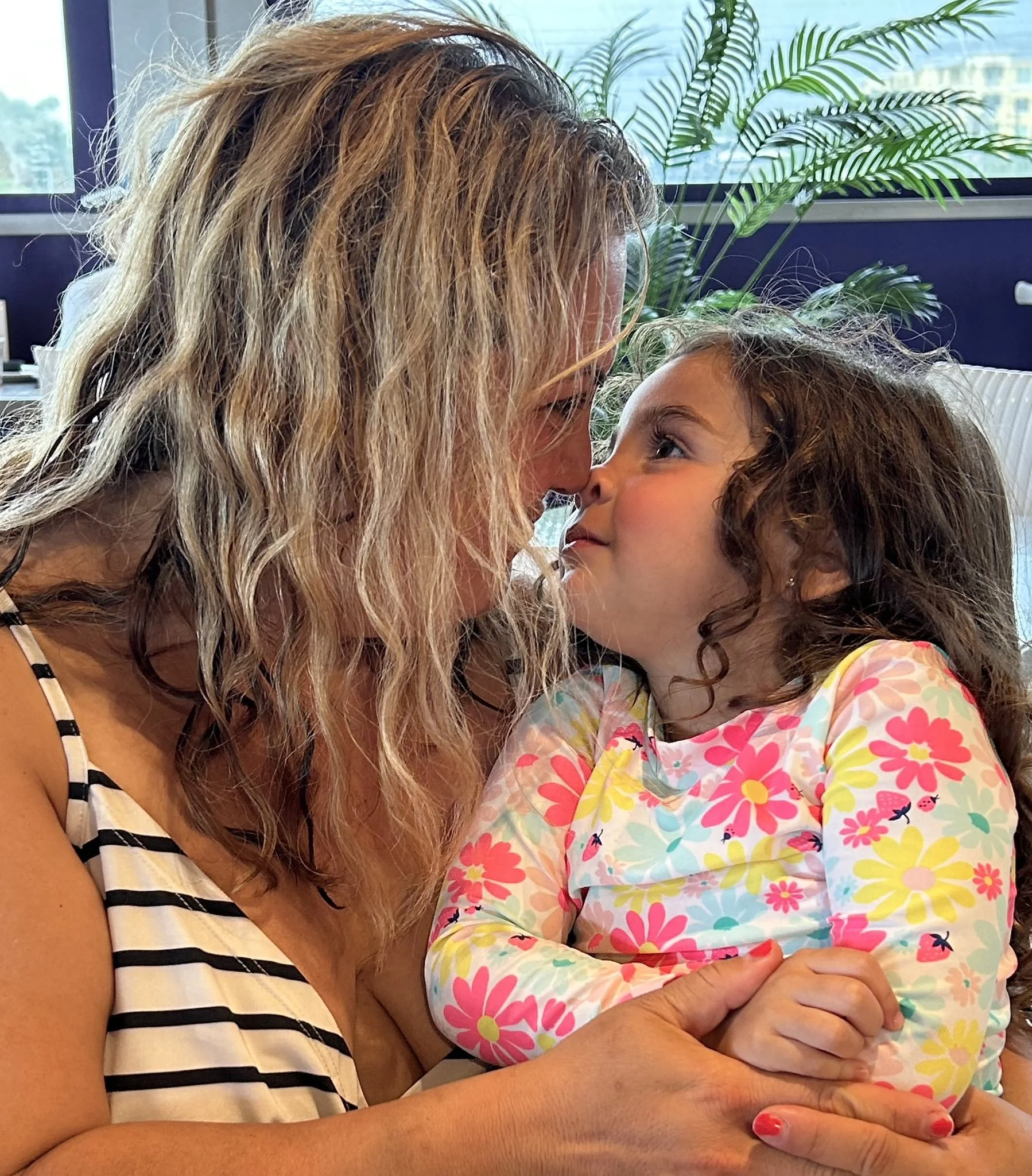 A woman with wavy blonde hair and a young girl with curly brown hair are sitting close together. The woman is wearing a black and white striped sleeveless top, and the girl is dressed in a colorful floral-patterned shirt. They are touching foreheads and looking into each other's eyes, with a large window and a green plant in the background.