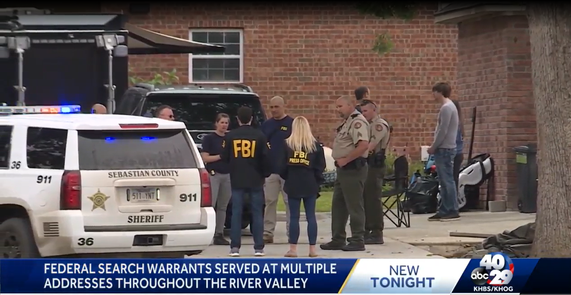 Law enforcement officers and FBI agents gathered outside a brick building, with marked sheriff and FBI vehicles nearby, discussing a case. A person is being questioned or spoken to by officials. The scene includes various bystanders and a folding chair, suggesting a police operation or investigation.