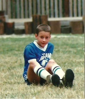 Young boy sitting on the grass in a sports uniform with socks and cleats, smiling at the camera.
