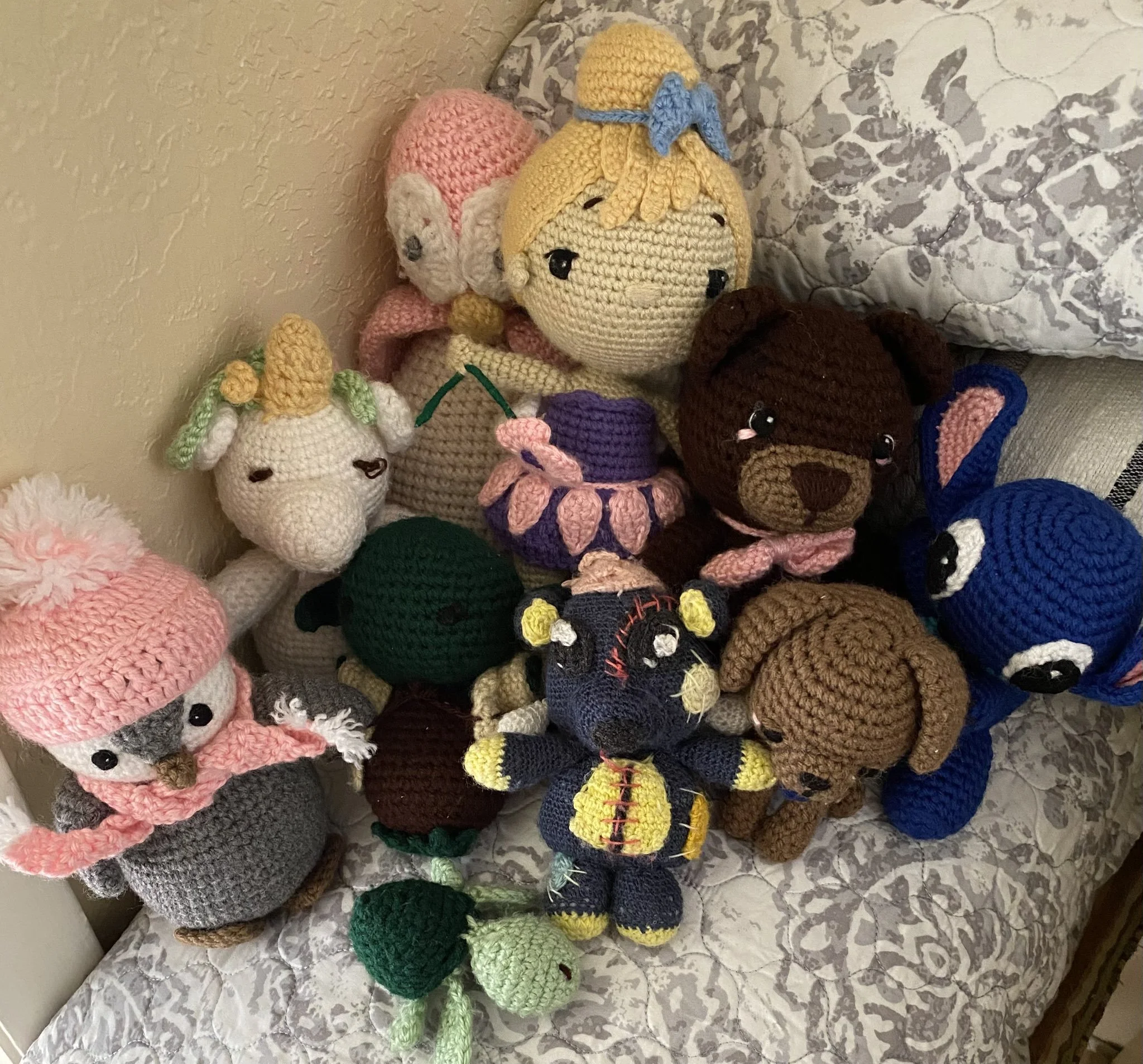 Collection of crocheted stuffed animals, including a pink hedgehog, a yellow character with a blue bow, a unicorn, a frog, and other animals, displayed on a fabric surface.