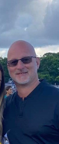 Bald man wearing sunglasses and a dark shirt, smiling outdoors with trees and cloudy sky in the background.