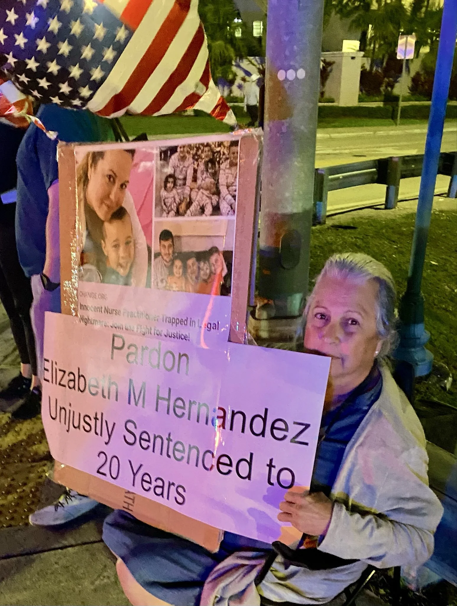 A person holding a poster with photographs and text, partially visible, about Elizabeth M. Hernandez, with an American flag balloon visible in the background. The scene appears to be outdoors at night.