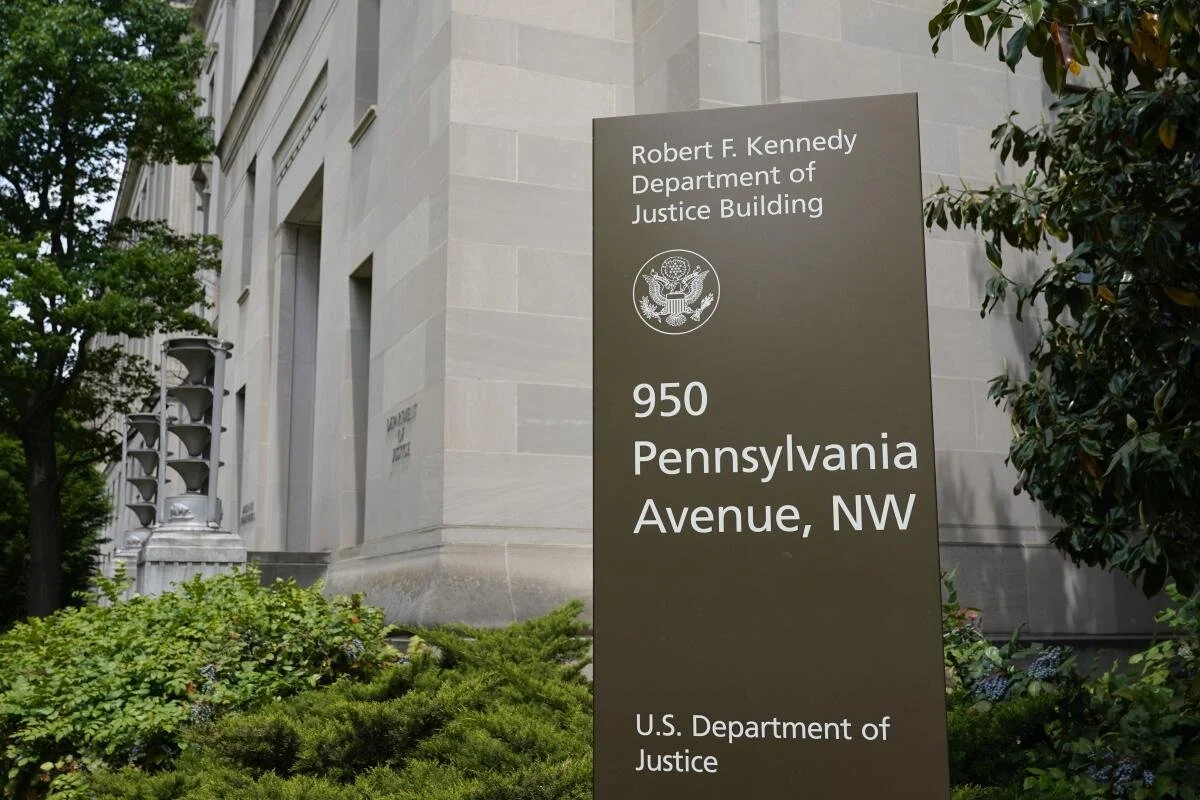 Signboard outside a government building reading 'Robert F. Kennedy Department of Justice Building, 950 Pennsylvania Avenue, NW, U.S. Department of Justice' with some greenery and part of the building visible in the background.