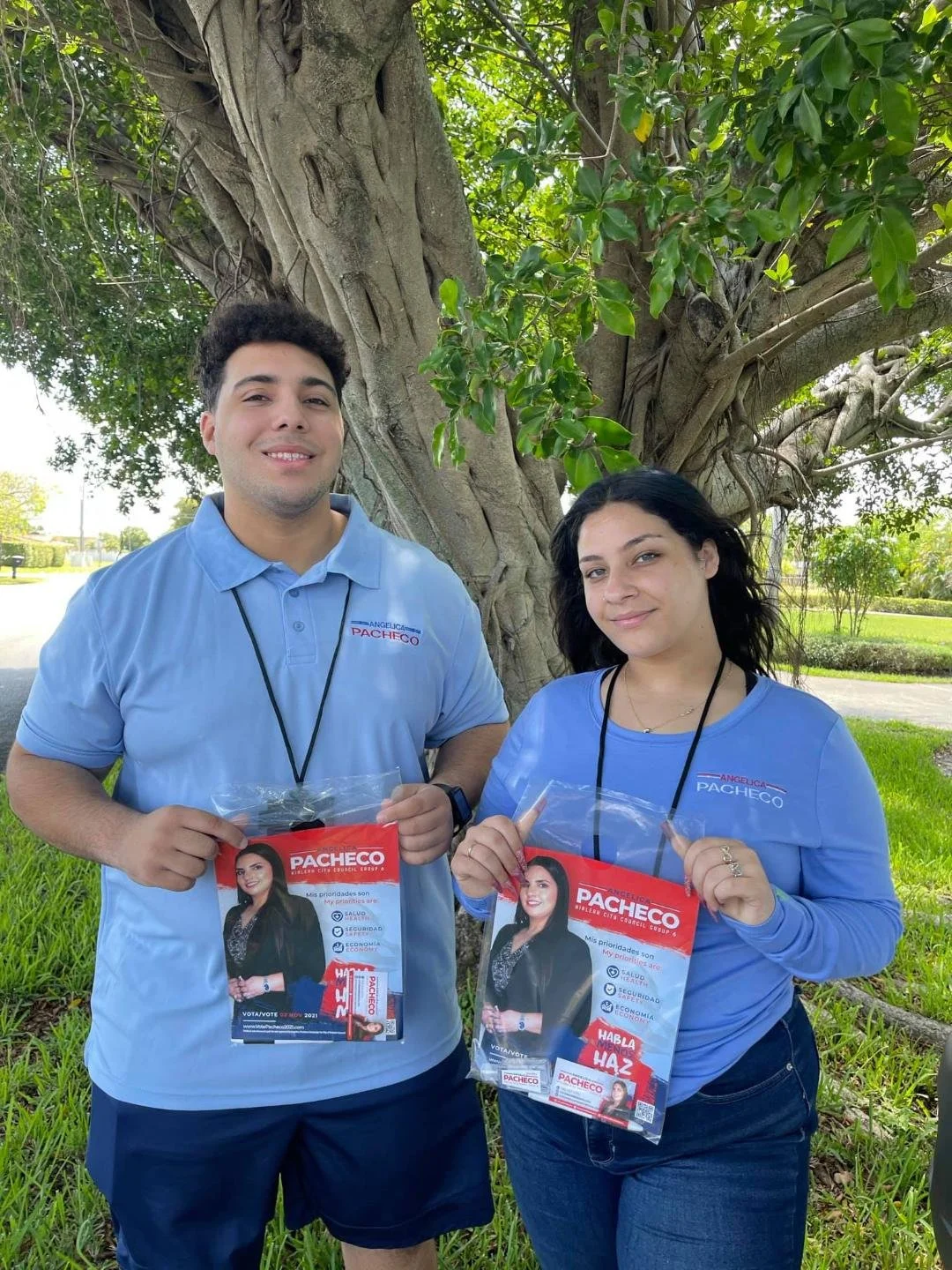 A young man and woman standing outdoors near a large tree, holding campaign flyers for Pacheco with a photo of a woman candidate. They are dressed in light blue shirts with Pacheco's logo.