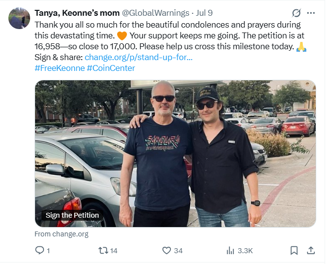 Two men standing outdoors in a parking lot, posing for a photo. One is wearing sunglasses and a graphic T-shirt, the other is wearing sunglasses, a hat, and a dark button-up shirt.
