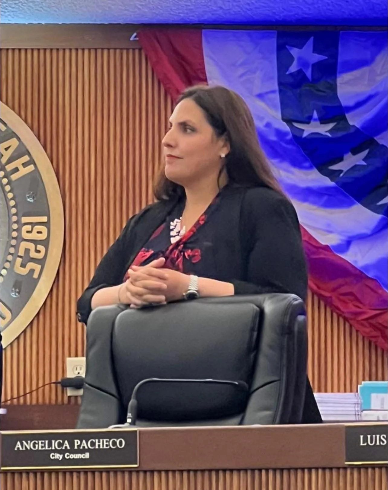A woman standing behind a desk at a city council meeting, with a nameplate that reads 'Angelica Pacheco, City Council'. She has dark hair, is wearing a black blazer, red patterned scarf, and has her hands clasped in front of her. Behind her, there is a Texas state flag and a wooden wall.
