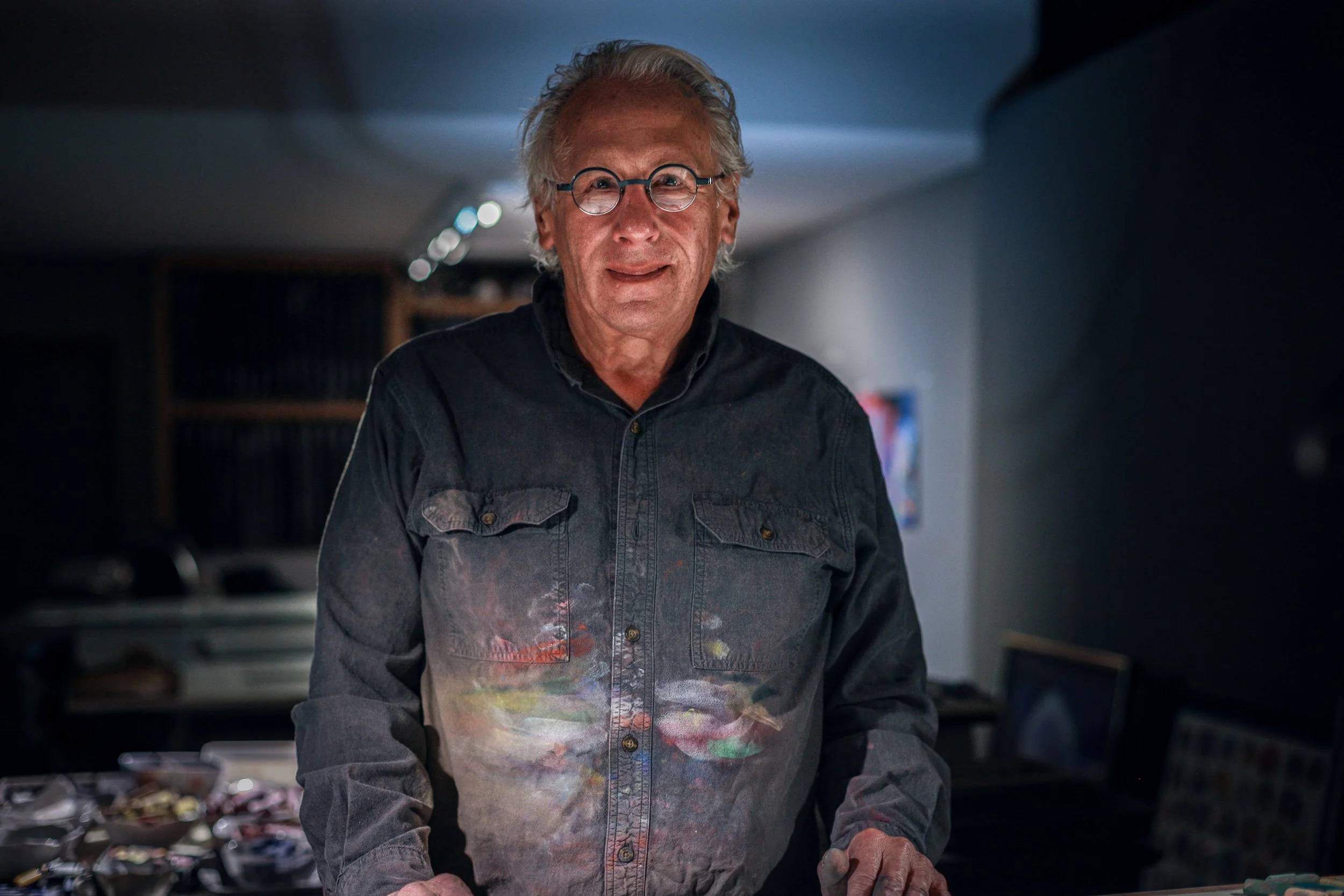 An elderly painter with gray hair and glasses standing in a dimly lit room, smiling, wearing a dark denim shirt with paint stains, surrounded by art supplies.