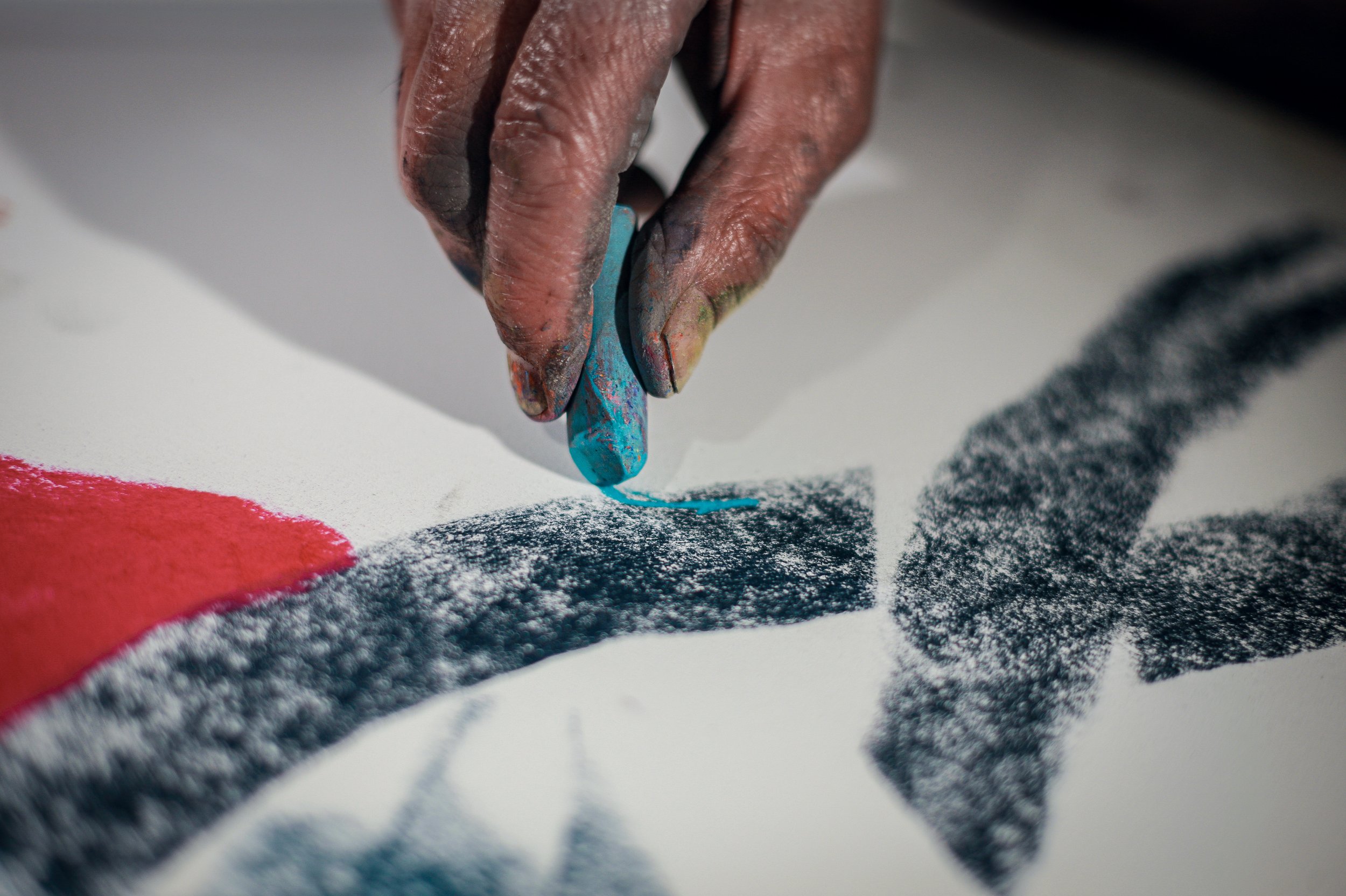 An artist holding a blue pastel is drawing on a large paper with colored pastels nearby.