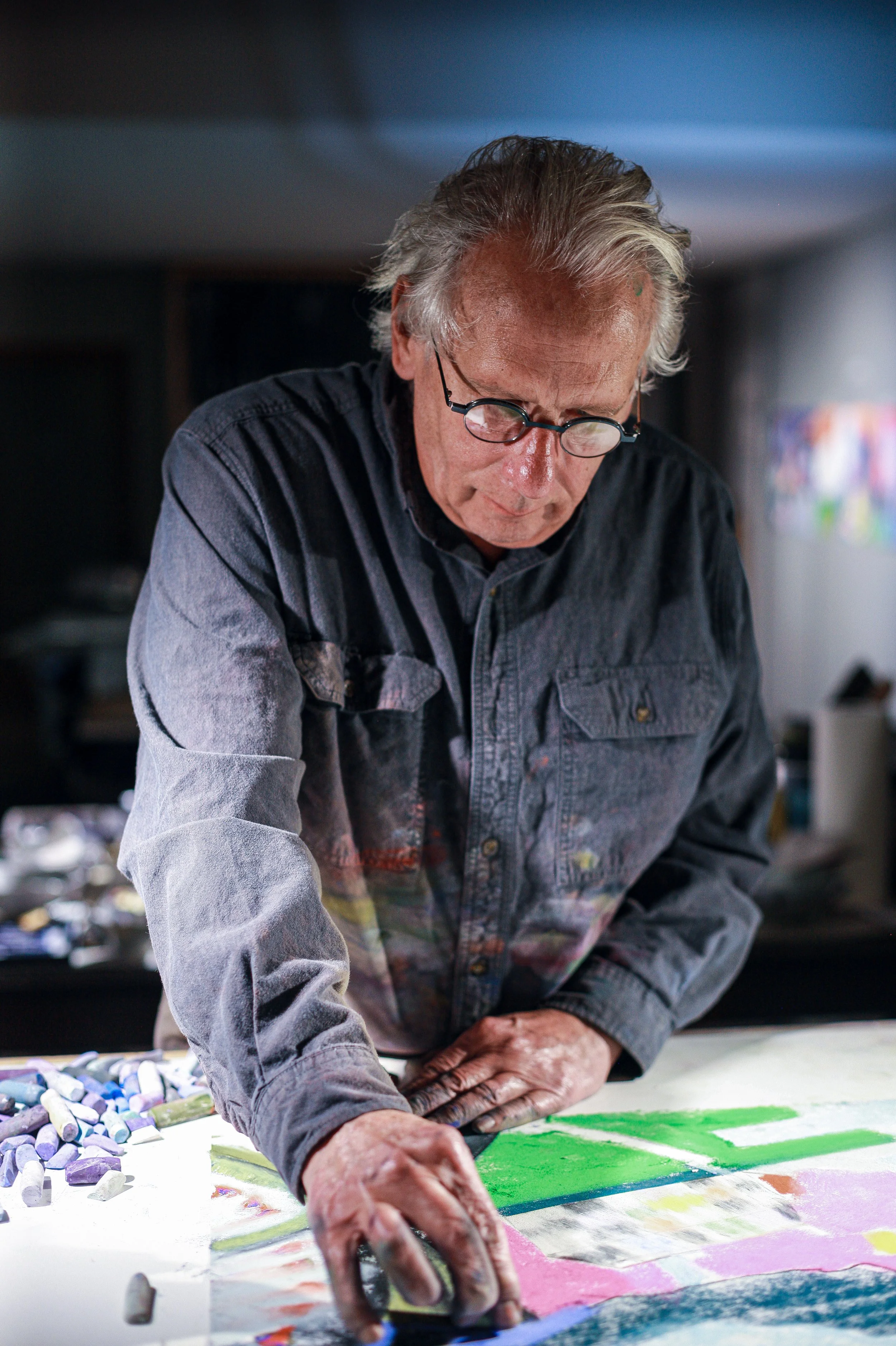 An older man with gray hair and glasses creating colorful artwork on a large sheet of paper with chalk or pastels, surrounded by art supplies.
