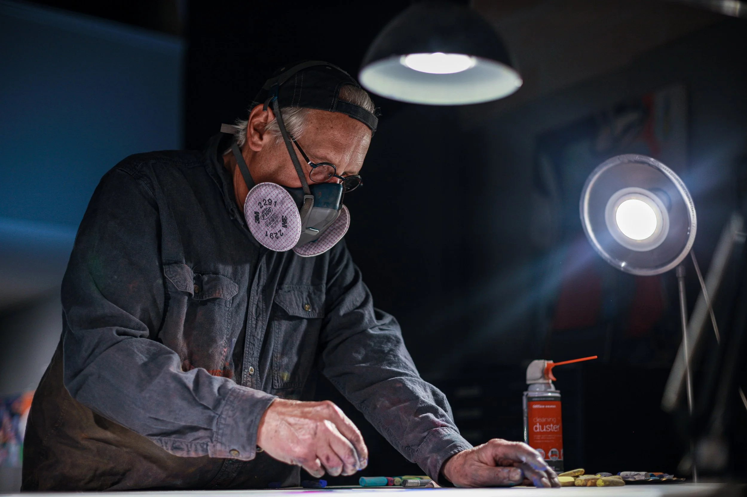 An older artist wearing protective mask and glasses working at a table with art supplies, illuminated by a bright overhead light in a workshop.