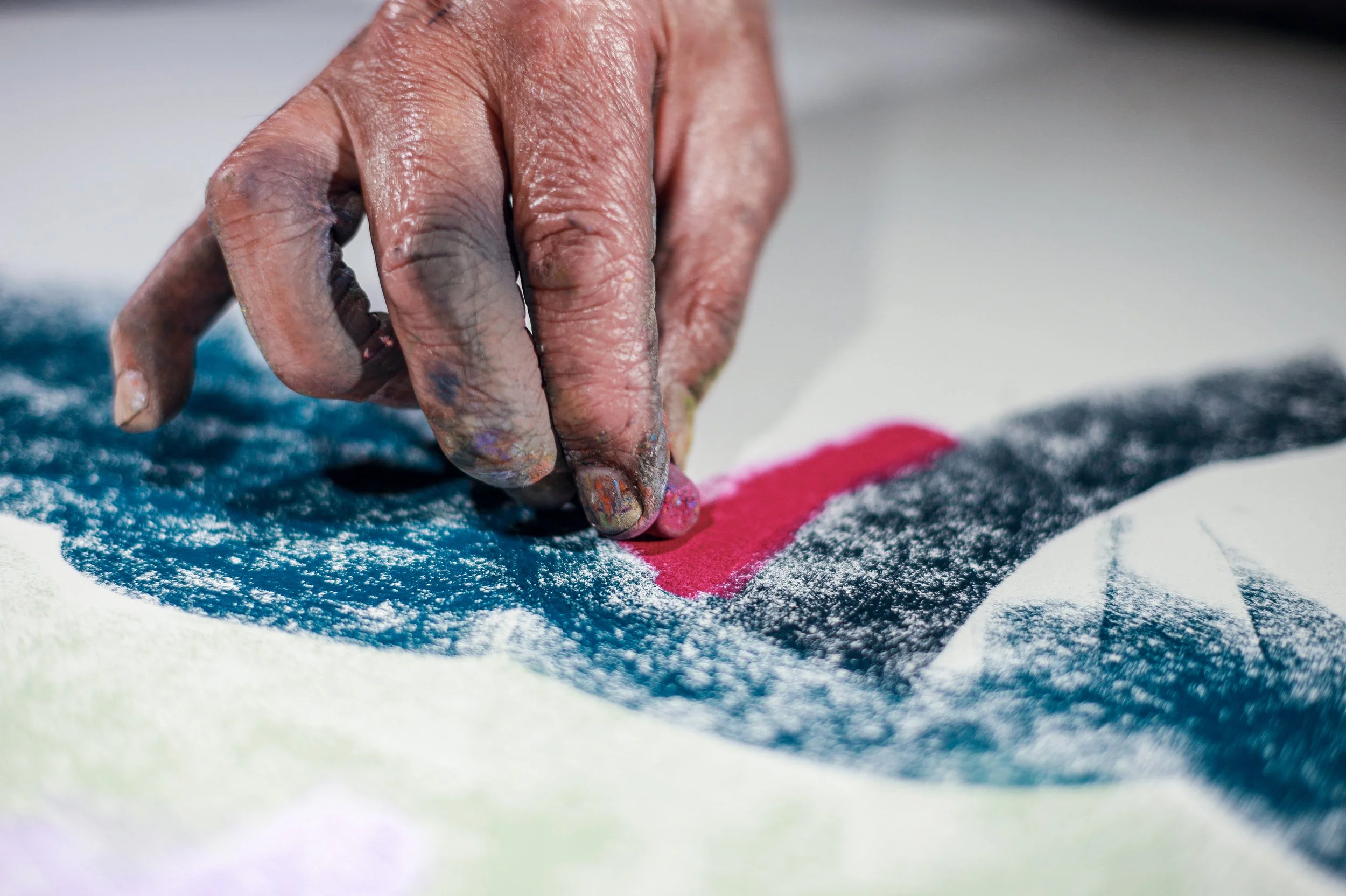 Close-up of a hand drawing on a textured surface with colorful pastels, including pink, blue, black, and white colors.