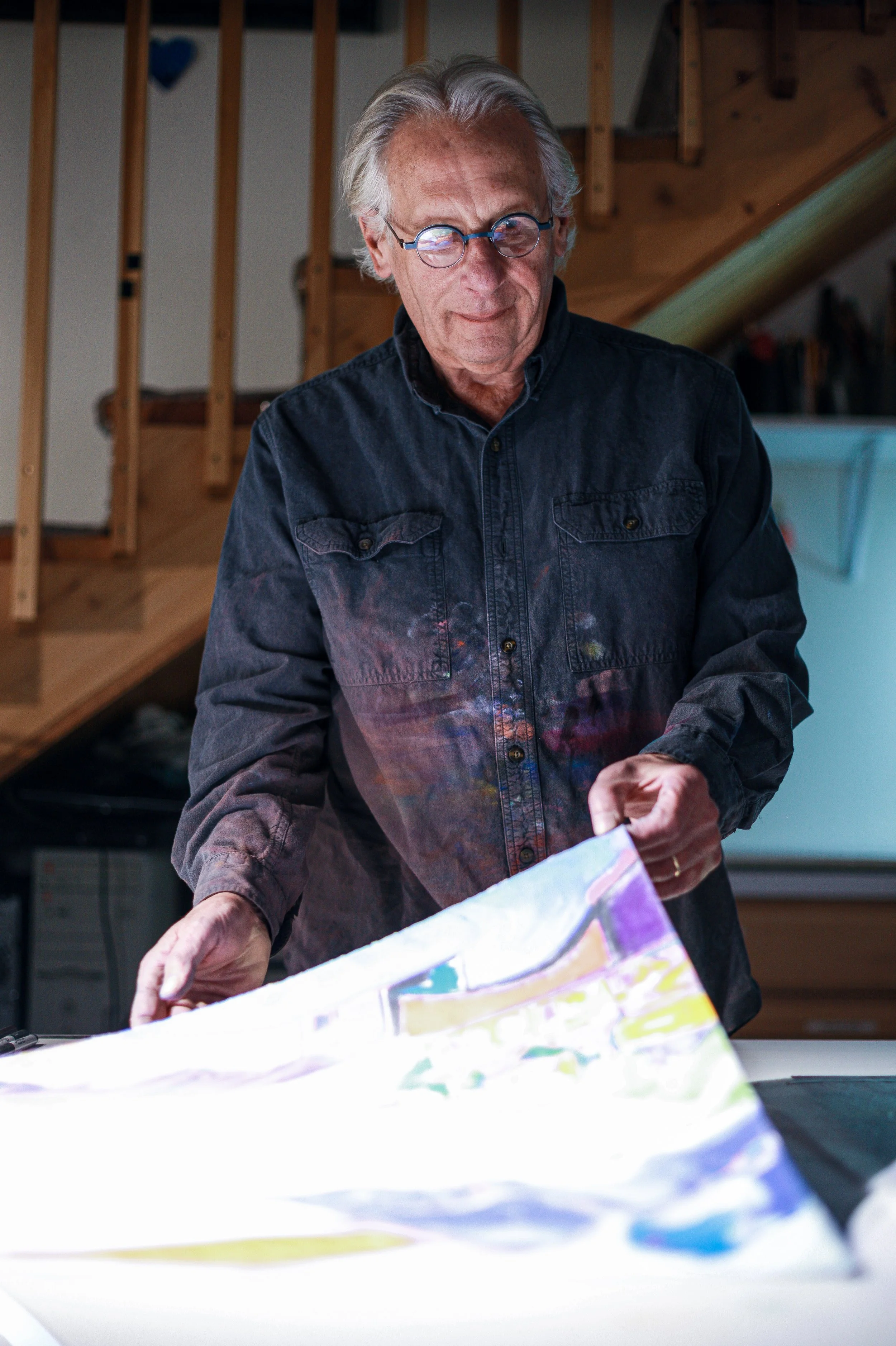 An older man with gray hair and glasses, wearing a dark, paint-splattered shirt, standing in a workshop or art studio, working on a colorful painting or art project.