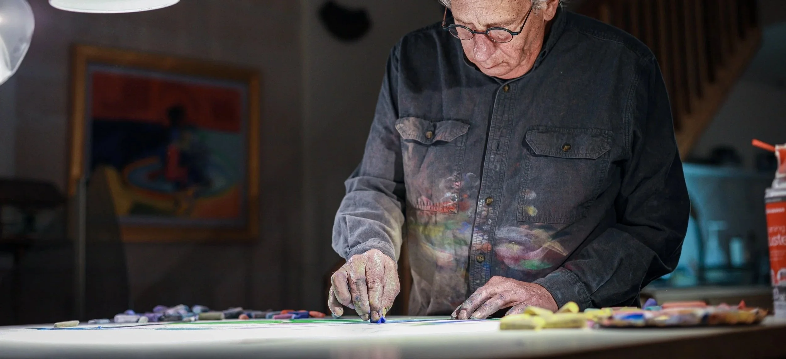 An elderly artist with glasses wearing a paint-stained denim shirt creating art with sidewalk chalk on a large white surface.