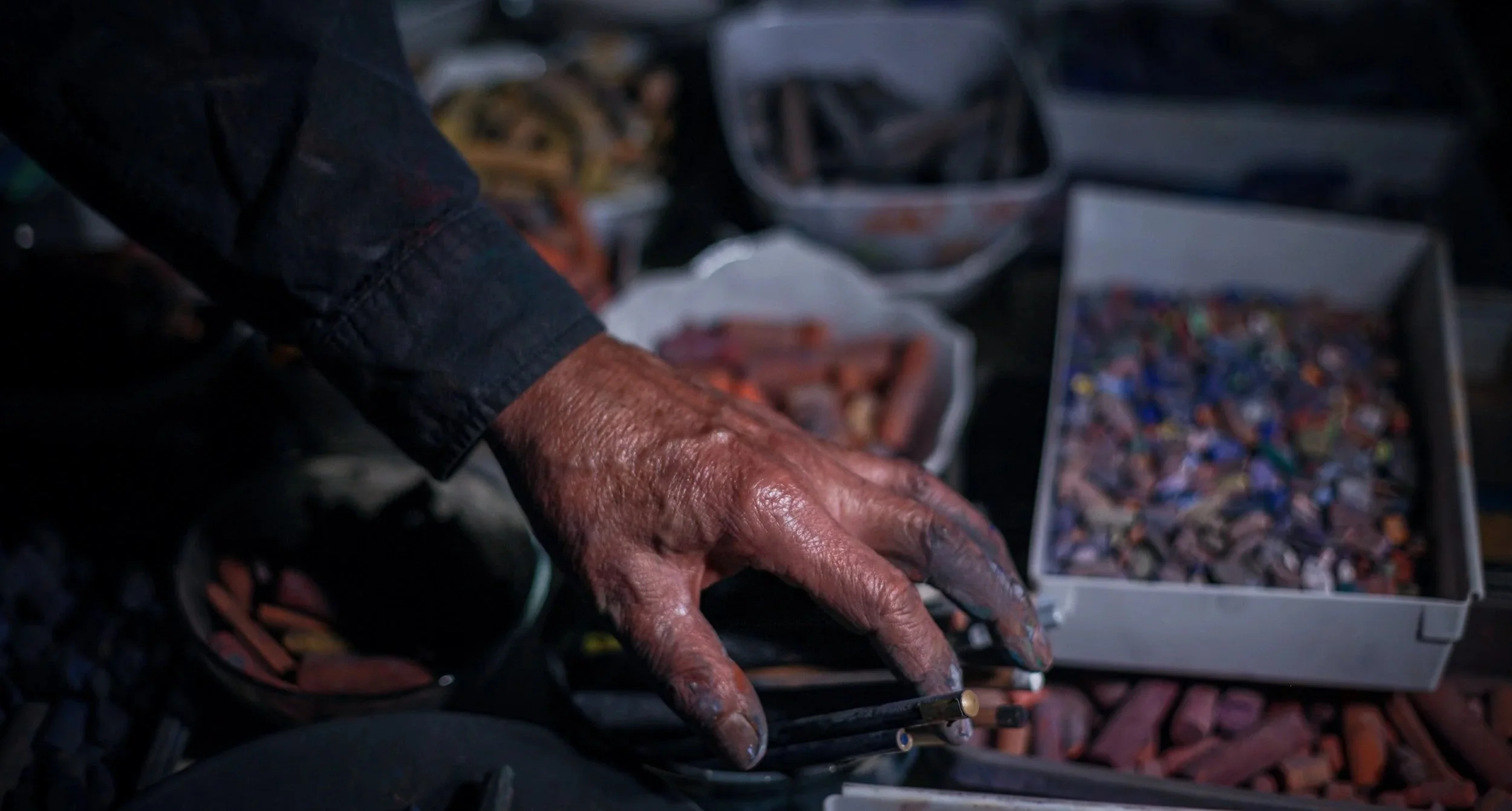 Close-up of a artist's hand reaching into an open box filled with soft pastels of various colors.