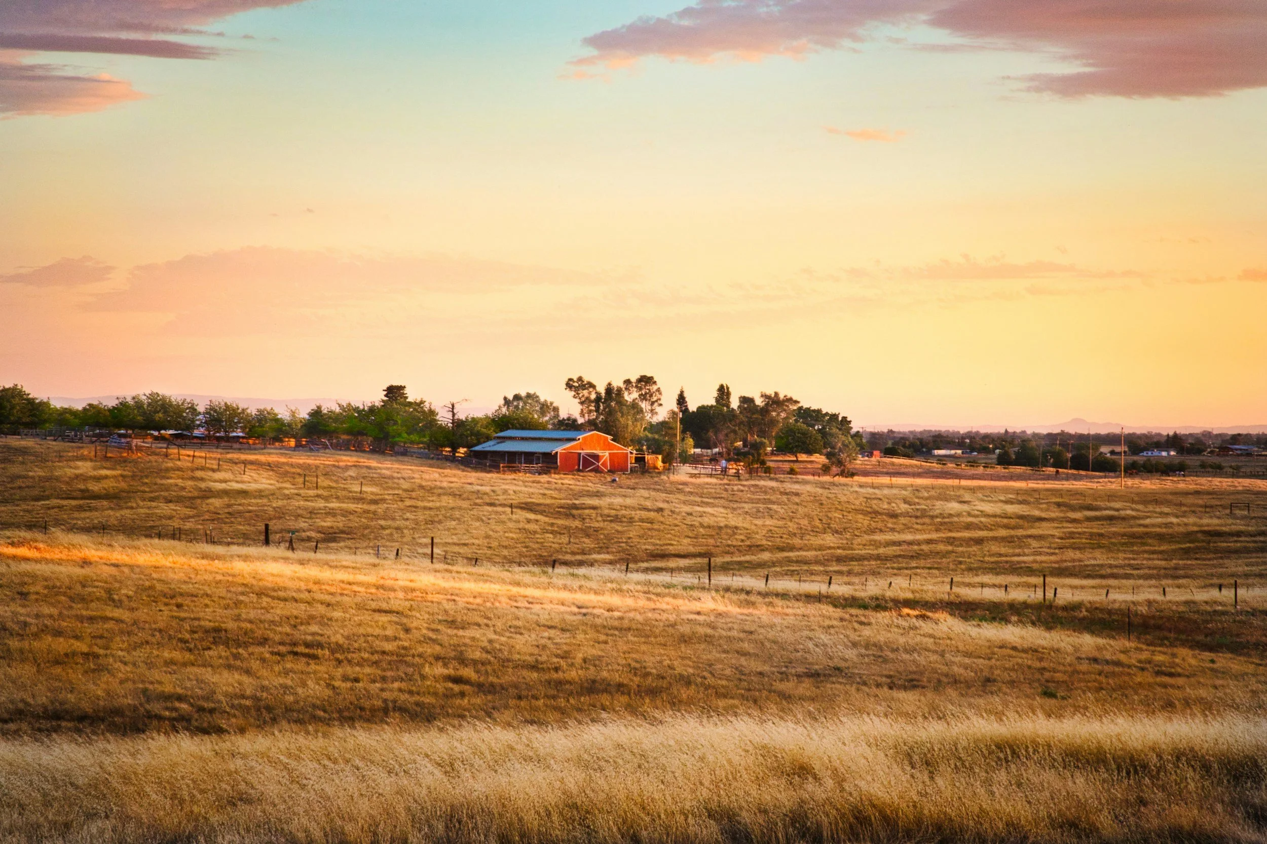 A rural landscape at sunset with a barn, trees, fencing, and open fields under a colorful sky with clouds.