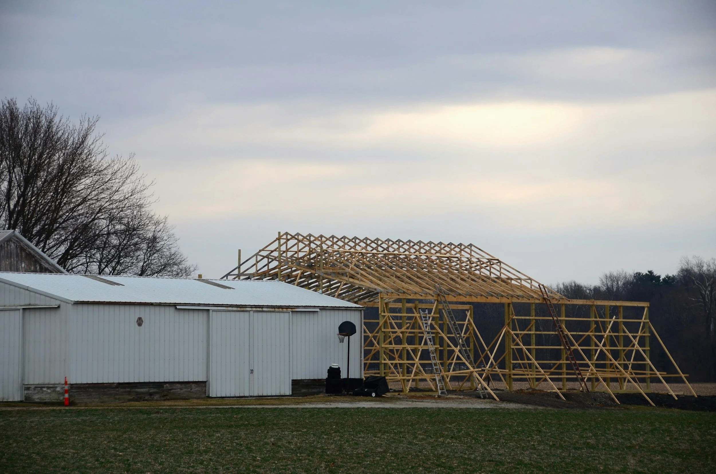 A house under construction with a wooden framework for the roof, a white metal shed in the foreground, and leafless trees under an cloudy sky.