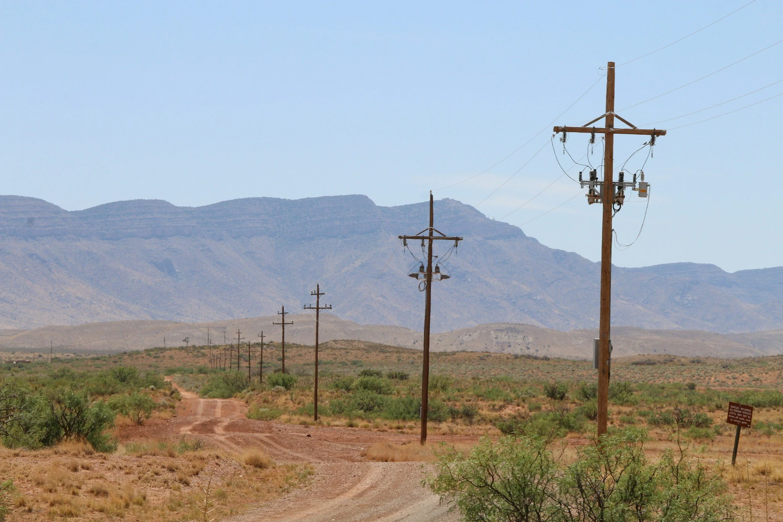 A rural landscape with a dirt road, multiple wooden utility poles with power lines, green bushes, mountains in the background, and clear blue sky.