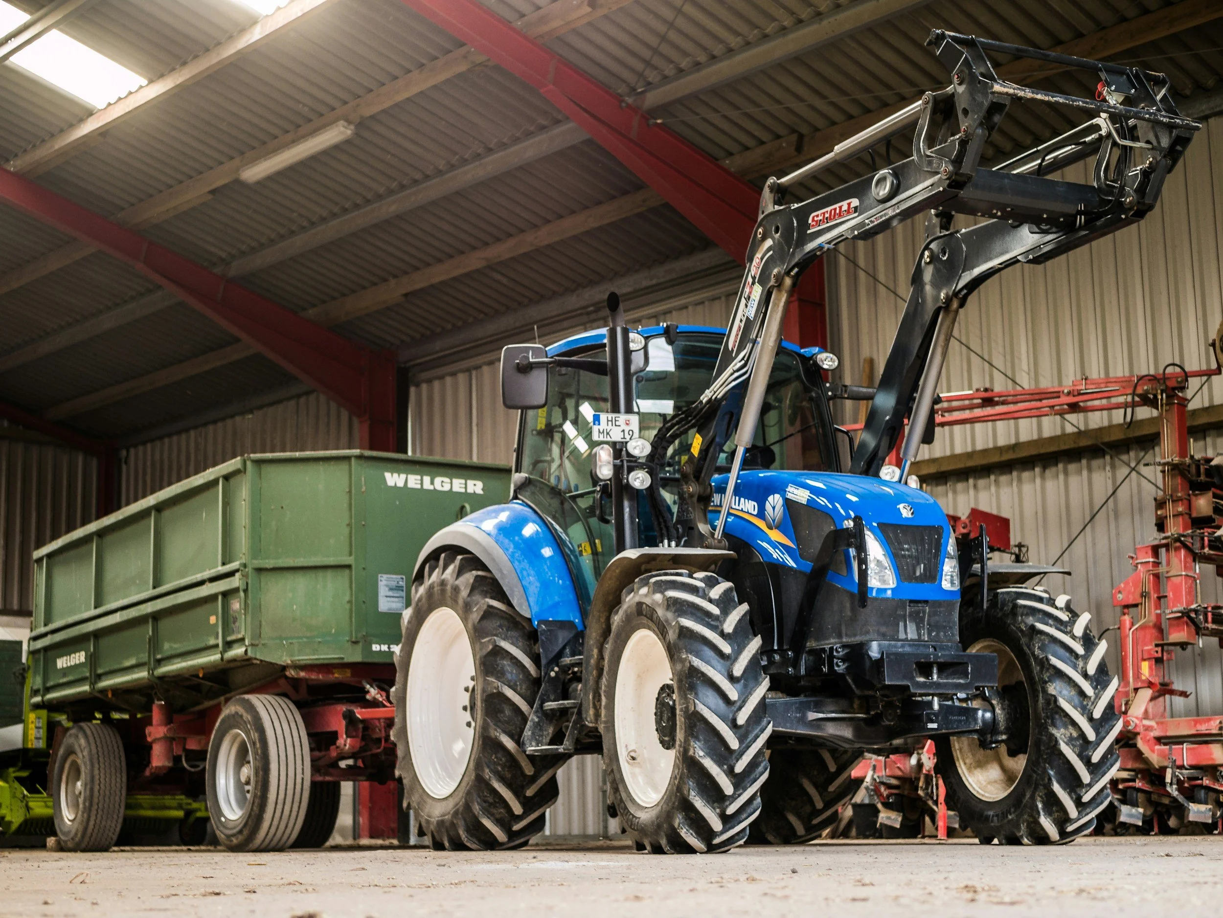 View of a tractor inside a storage warehouse, with a green metal trailer labeled "Welger" attached to it.