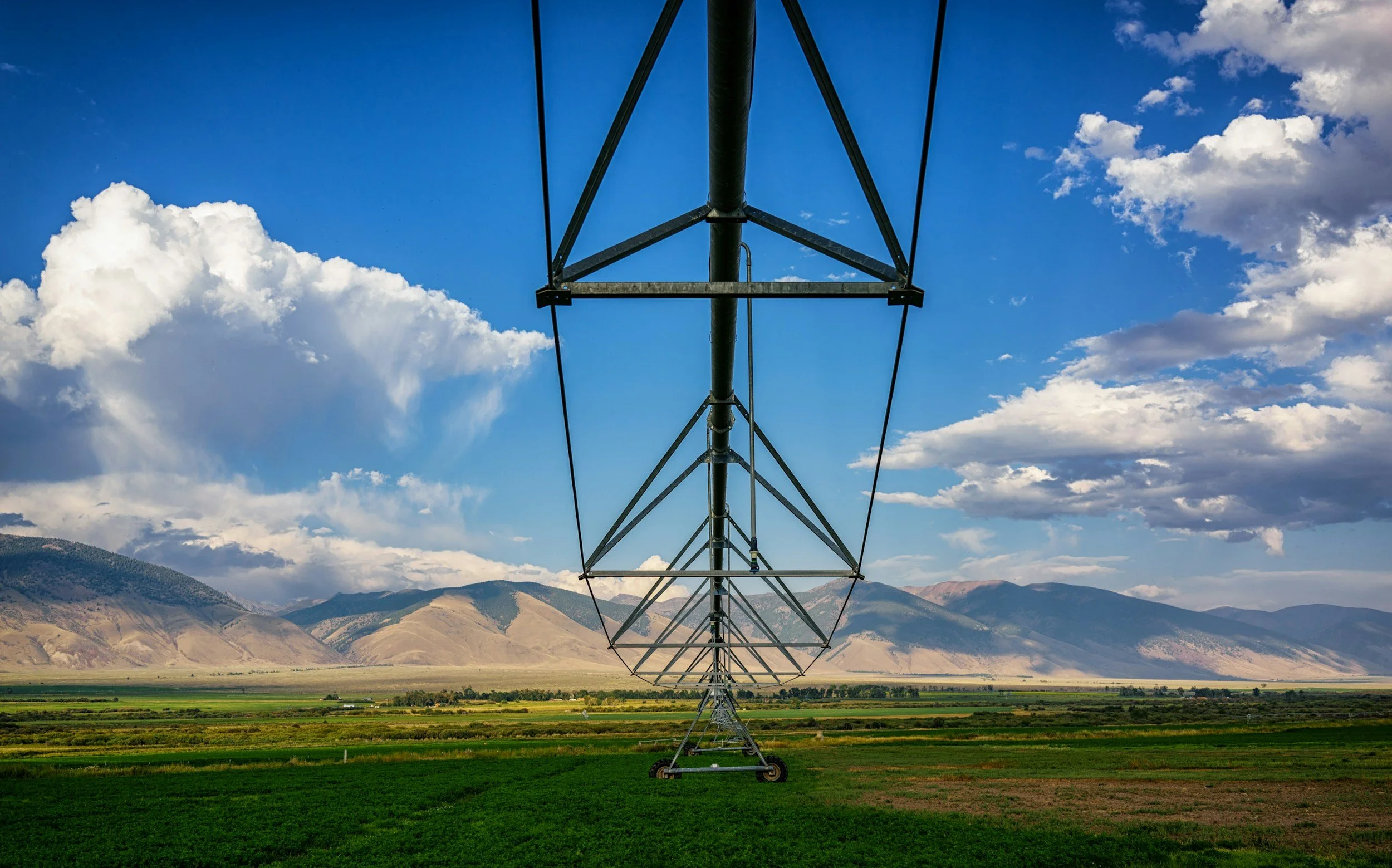 A large irrigation pivot system with metal framework extends across a green field, set against a backdrop of mountains and a partly cloudy sky.