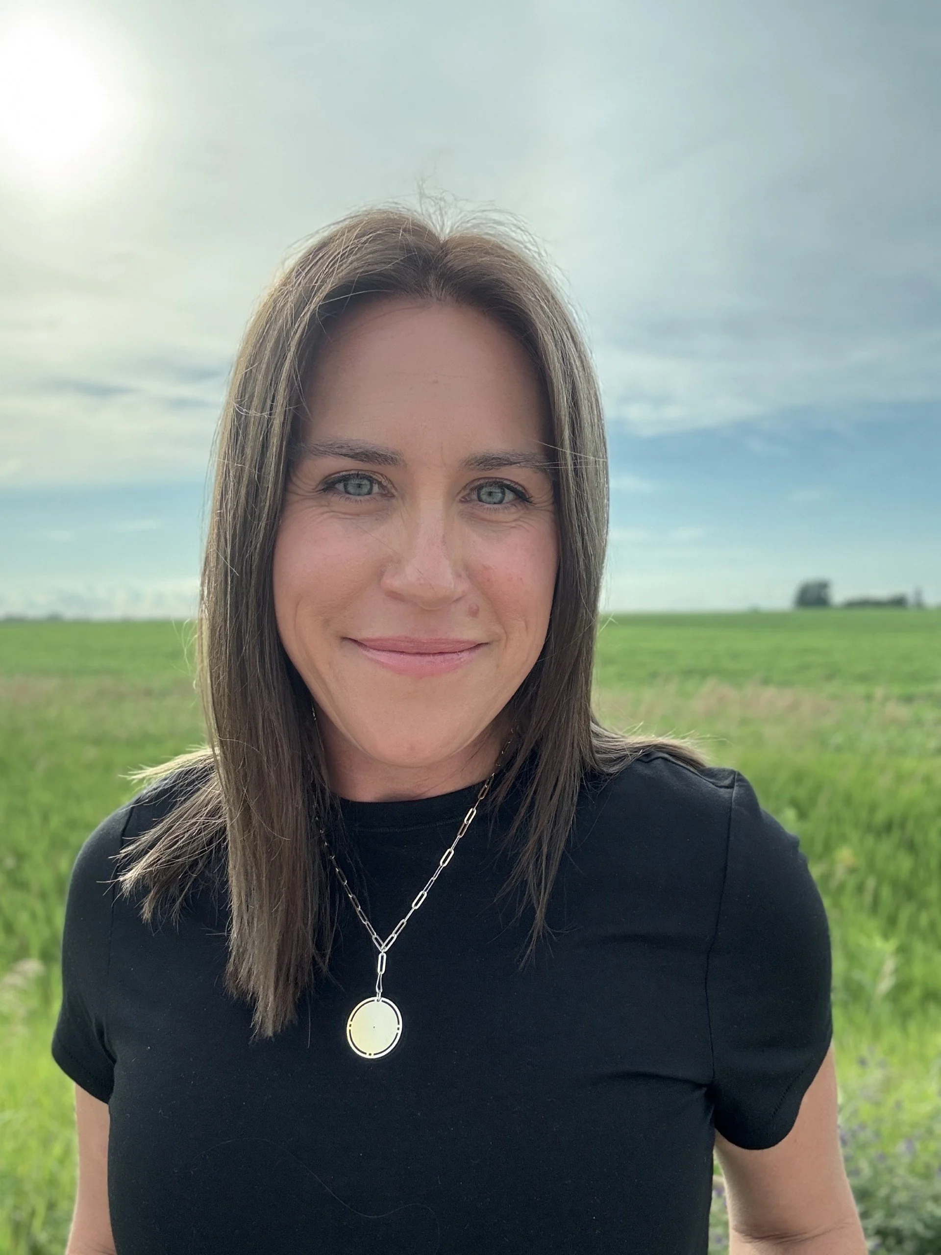 Close-up of a smiling woman with long brown hair, blue eyes, wearing a black top and a silver necklace with a round pendant, standing outdoors in a green field with a partly cloudy sky in the background.