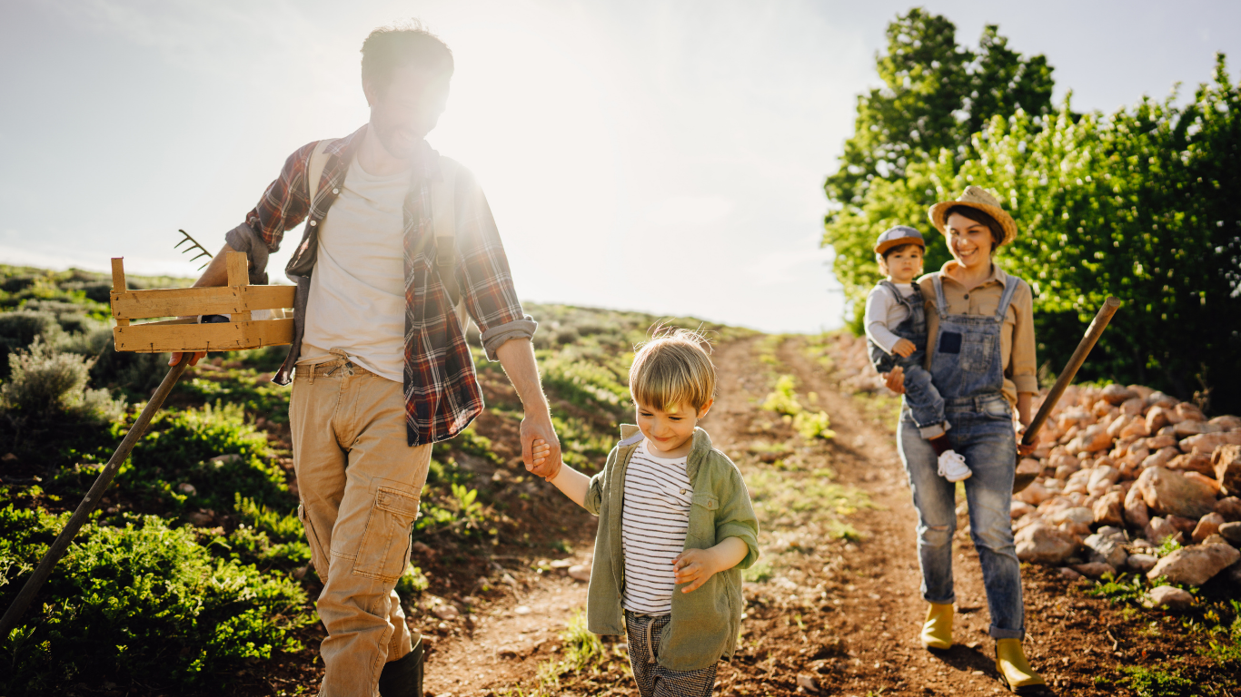 Family hiking outdoors on a sunny day with a father, mother, and two children, walking on a dirt trail through greenery, with the father holding hands with a young boy and the mother carrying a toddler.