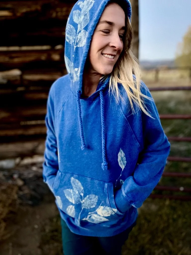 A woman smiling outdoors wearing a blue hoodie with white leaf patterns, hands in pockets, with a blurred wooden structure and fence in the background.