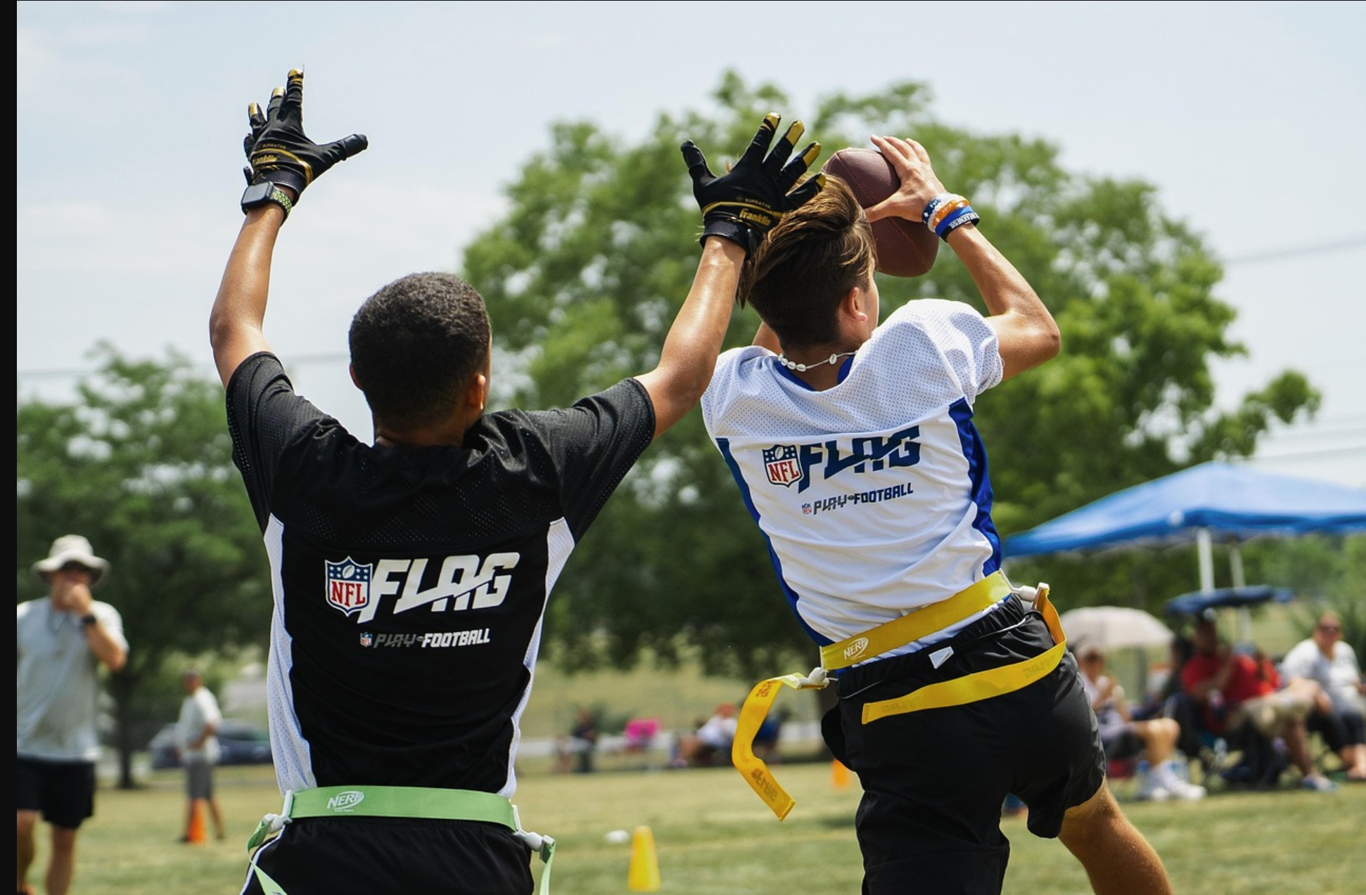 Two football players in uniforms compete for a football in an outdoor game. One player is jumping to catch the ball, while the other has their hands raised. Spectators are seen in the background.