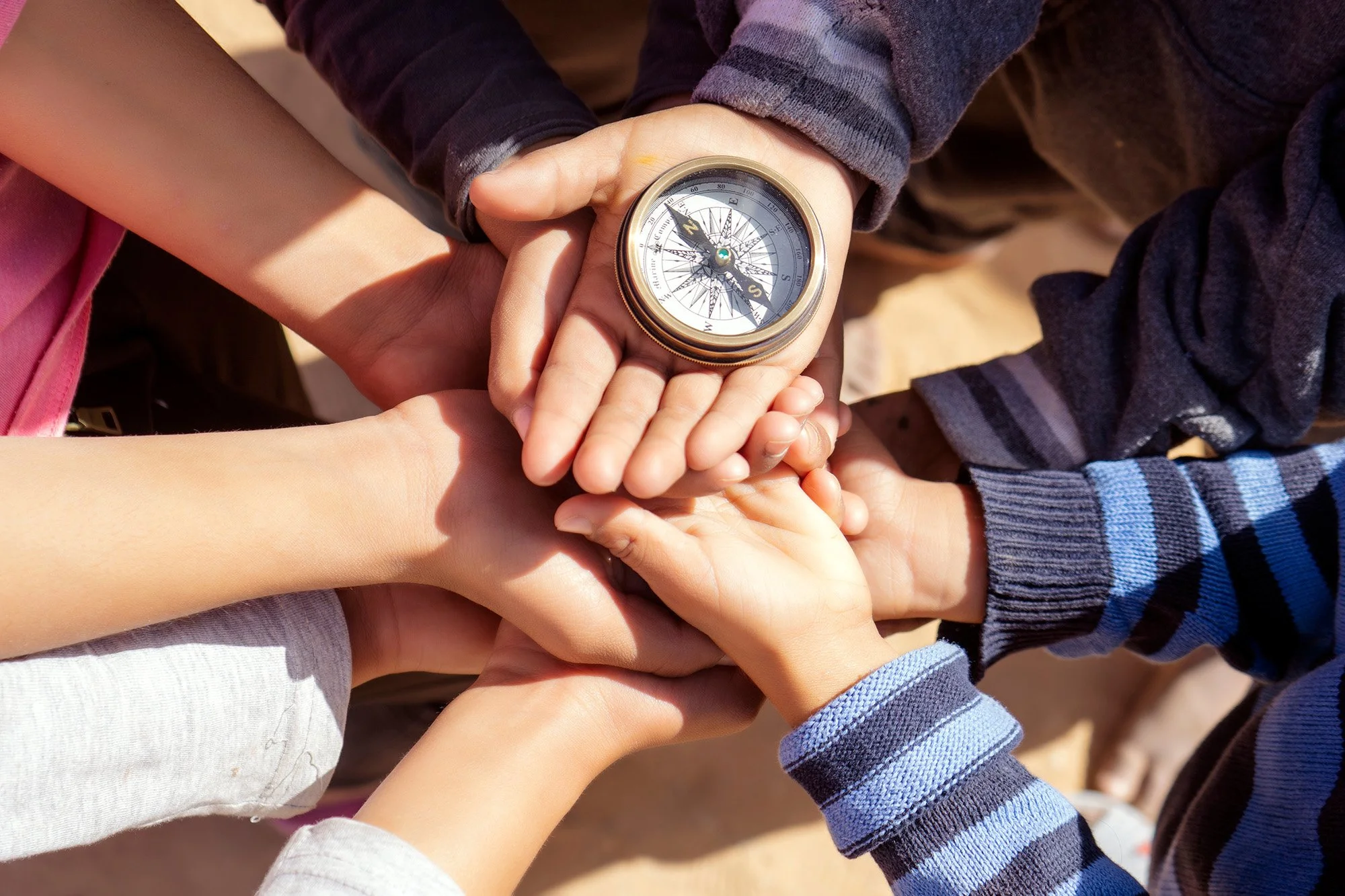 A group of children and one adult hands are stacked together, with the adult holding a compass. The setting appears to be outdoors on a sandy surface.