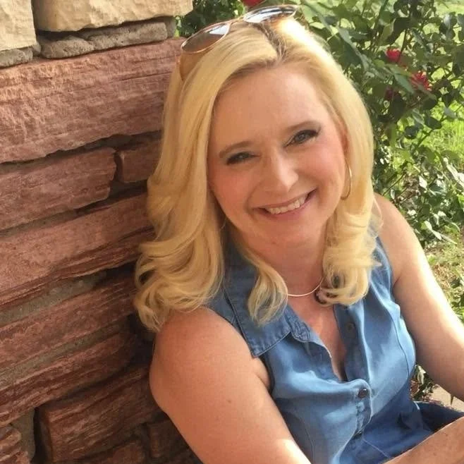 A blonde woman smiling while sitting outdoors near a brick wall and greenery.