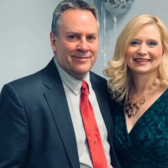 A man in a dark suit, light blue shirt, and red tie standing next to a woman with blonde hair wearing a dark teal dress with a necklace, both smiling indoors.