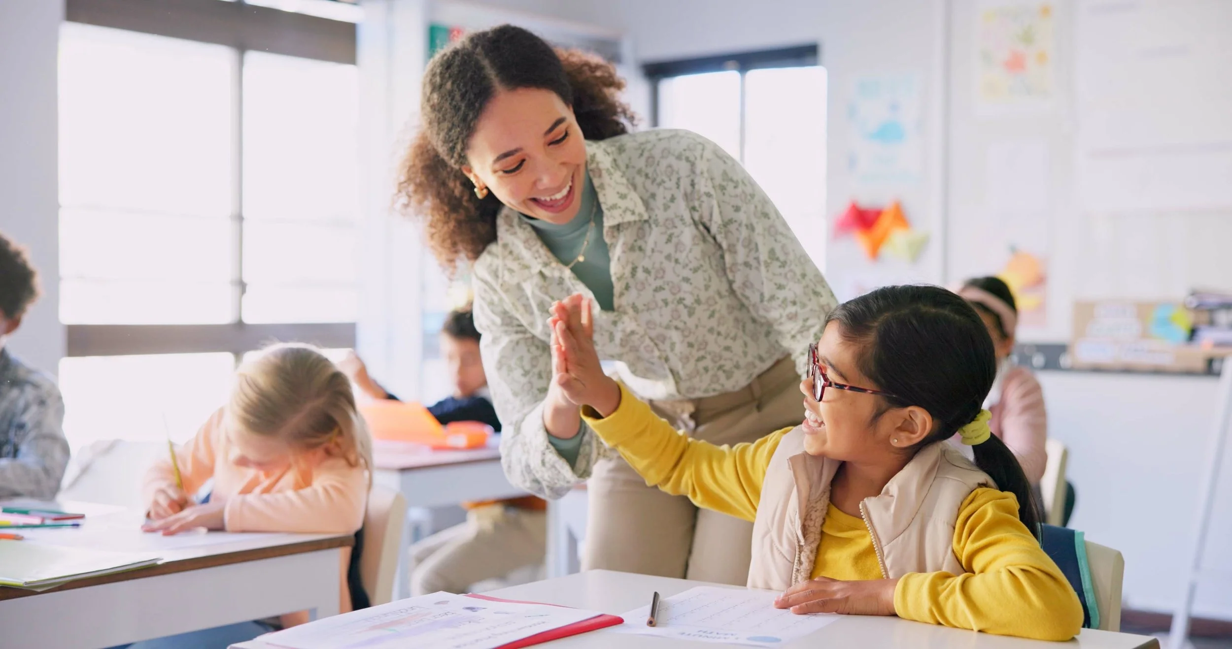 A teacher high-fives a student sitting at her desk in a classroom with other students.