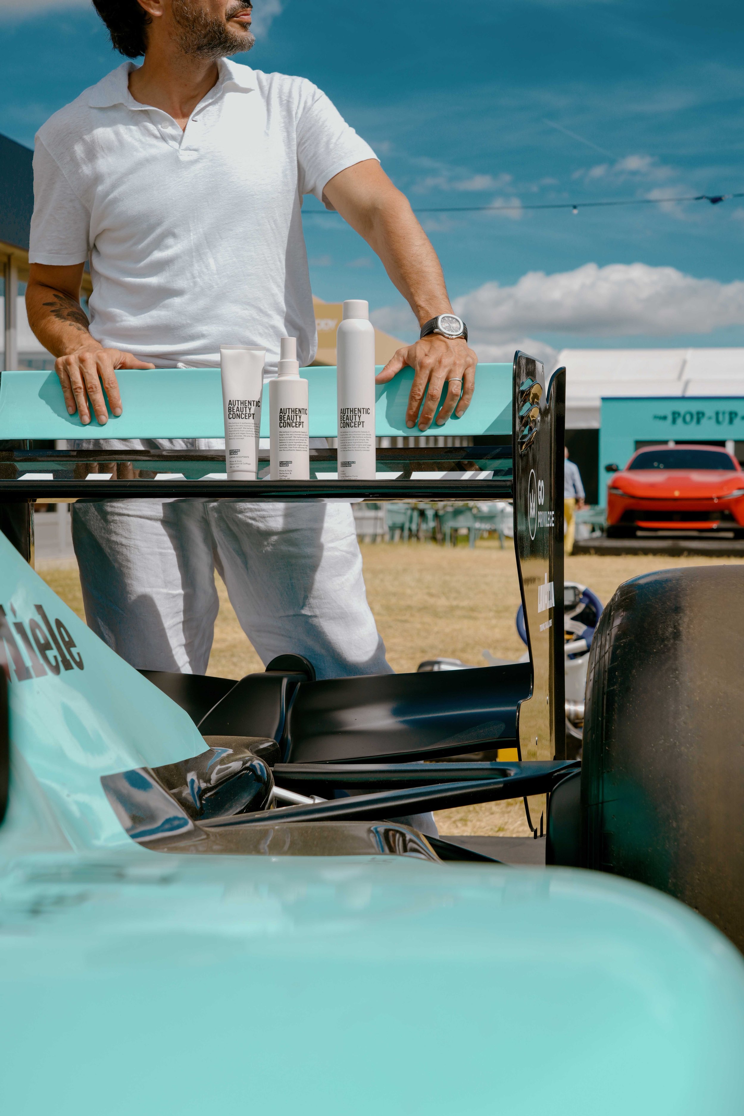 A man with gray hair and a beard, wearing a white polo shirt and white pants, standing next to a teal and black race car at an outdoor event on a sunny day with blue sky and clouds. The man has his hand on the car's rear wing, displaying skincare pro