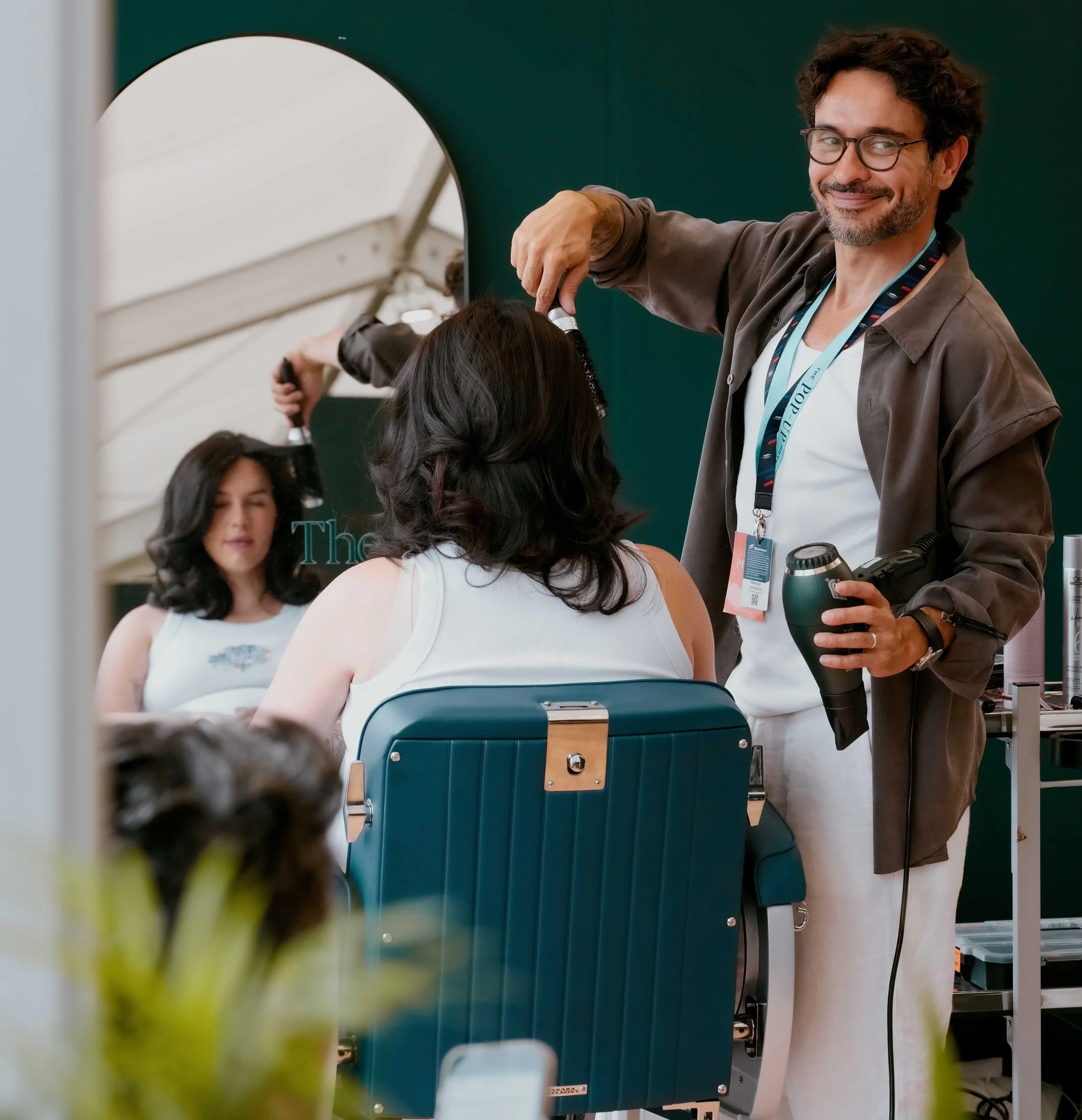 A smiling man with glasses and a beard holds a hairdryer, styling a woman's hair at a salon or beauty event. Other women are seated nearby, with hair and beauty equipment visible.