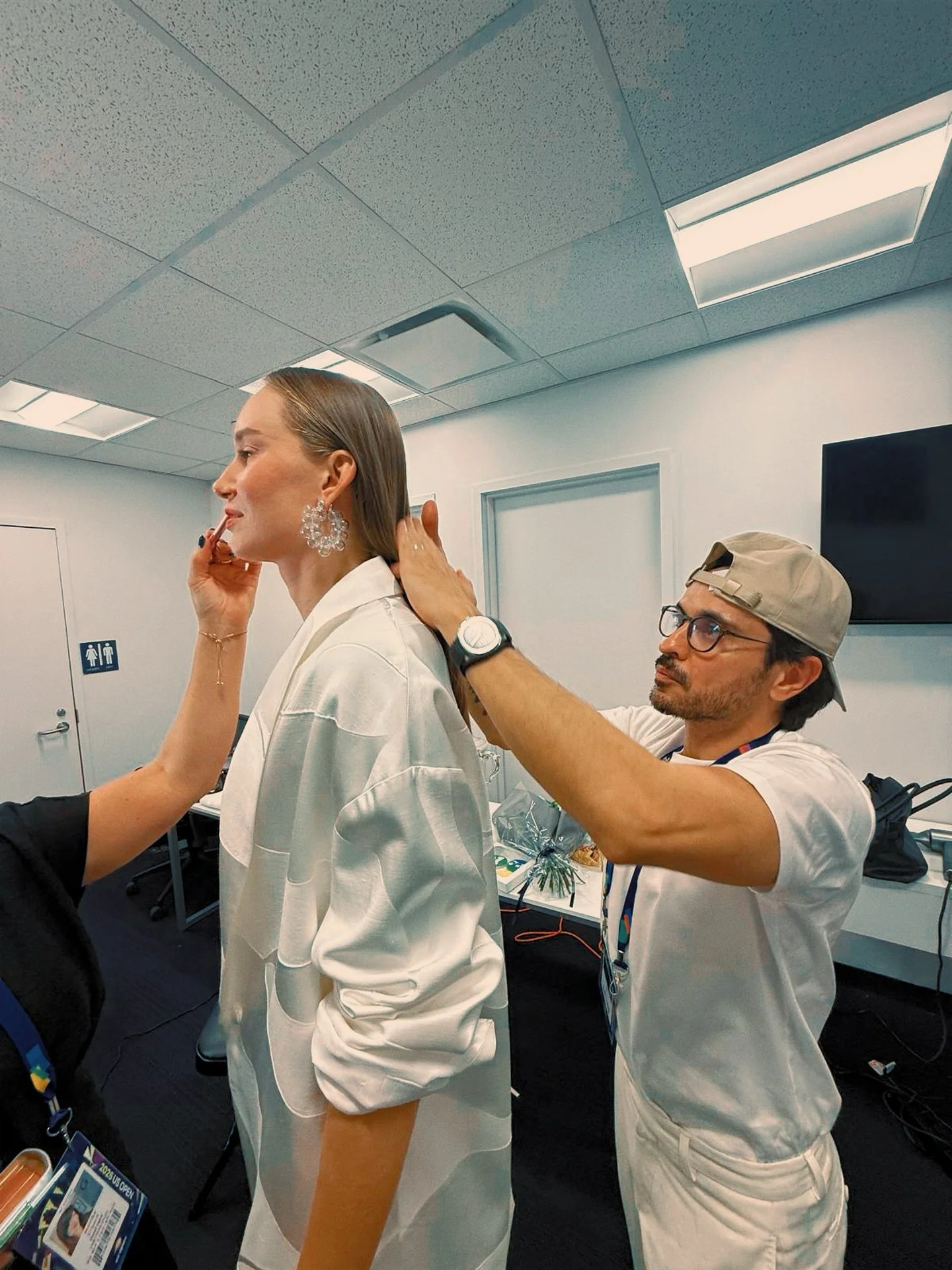 A woman with short hair wearing large earrings and a white blazer while another person helps adjust her hair in a room with a ceiling-mounted light and a television screen.