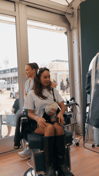 A woman sitting in a salon chair at an airport terminal, holding a paper bag, with another woman standing behind her. There are large glass windows and people visible outside.