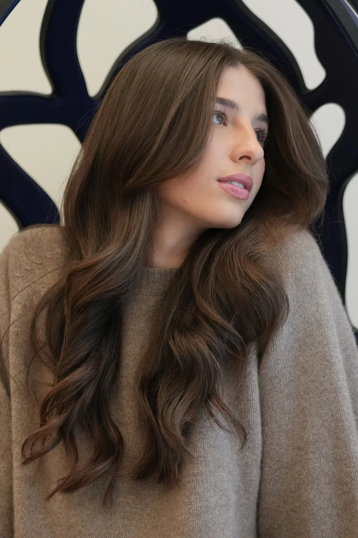 A woman with long, wavy brunette hair and blue eyes, wearing a beige sweater, sitting in front of an abstract black and white background.