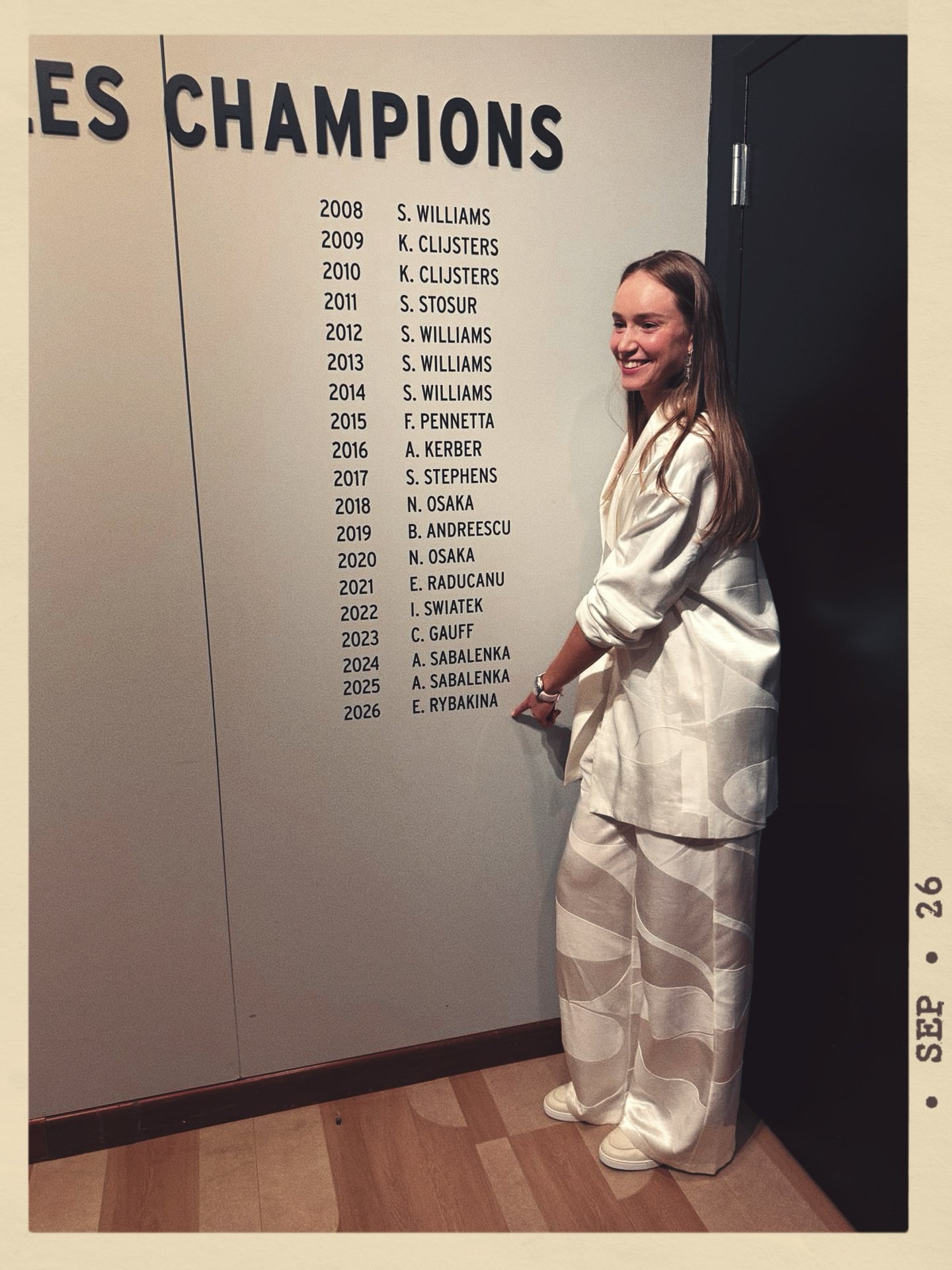 Elena Rybakina in a cream-colored suit standing next to a list of the US Open tennis champions on a wall, smiling.