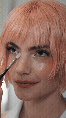 Woman with pink hair applying makeup with a brush near her eye.