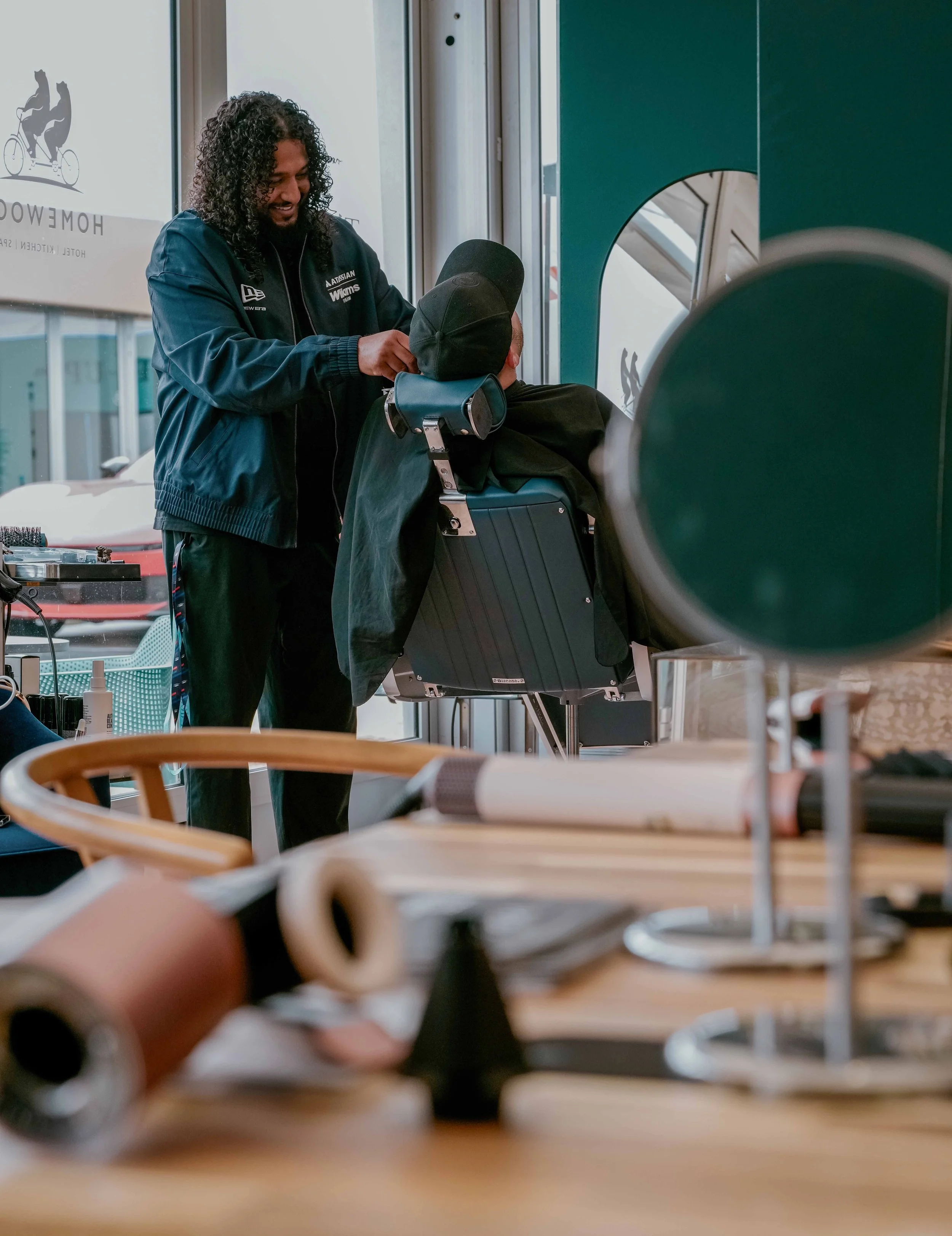 A man with curly hair and a beard, wearing a navy jacket, is helping a child in a wheelchair at a reception desk in an indoor setting. The child is wearing a cap and has a black jacket on. There are various items like notebooks and a telephone on the