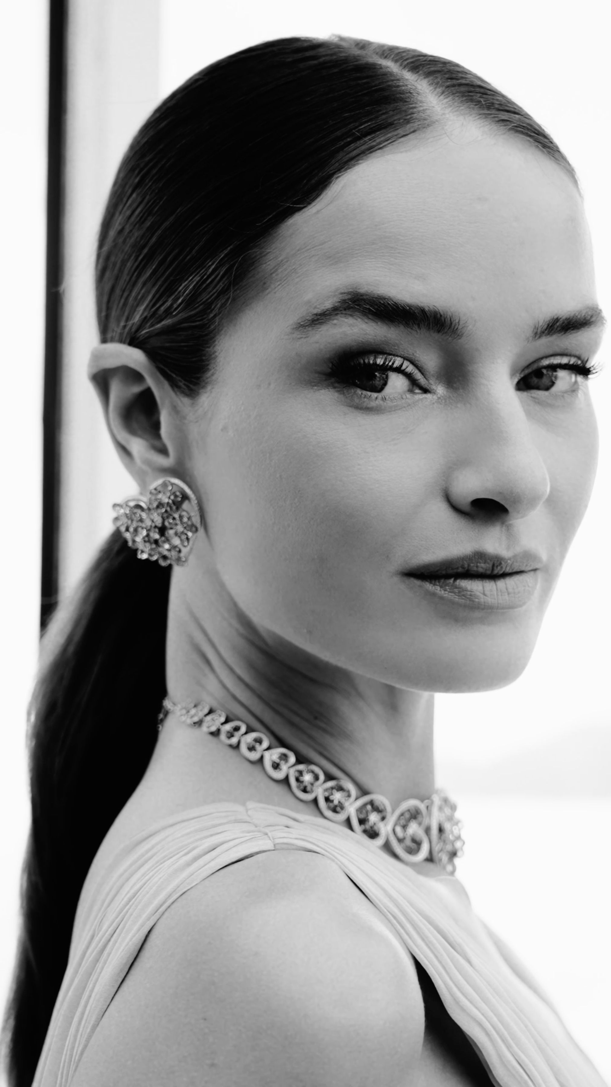 Black and white portrait of a woman with smooth, slicked-back hair, wearing large ornate earrings, a necklace, and a dress with pleats on the sleeve, looking over her shoulder with a neutral expression.