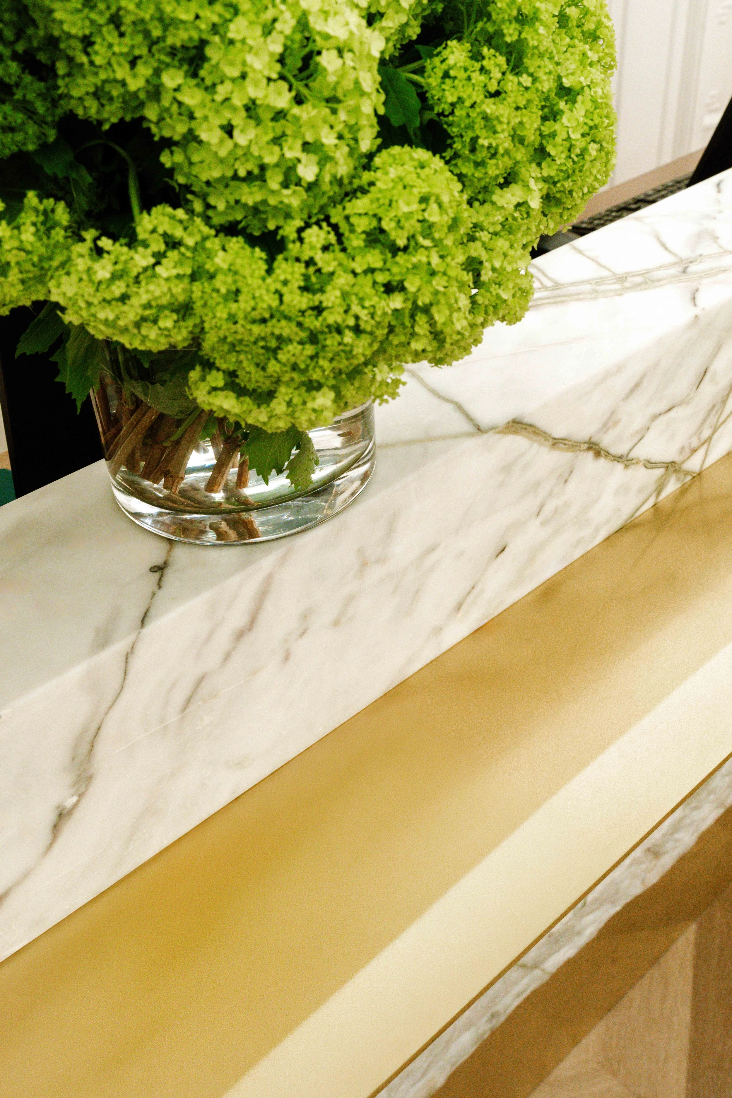 Close-up of a clear glass vase with green hydrangea flowers placed on a white marble surface, with a beige background.