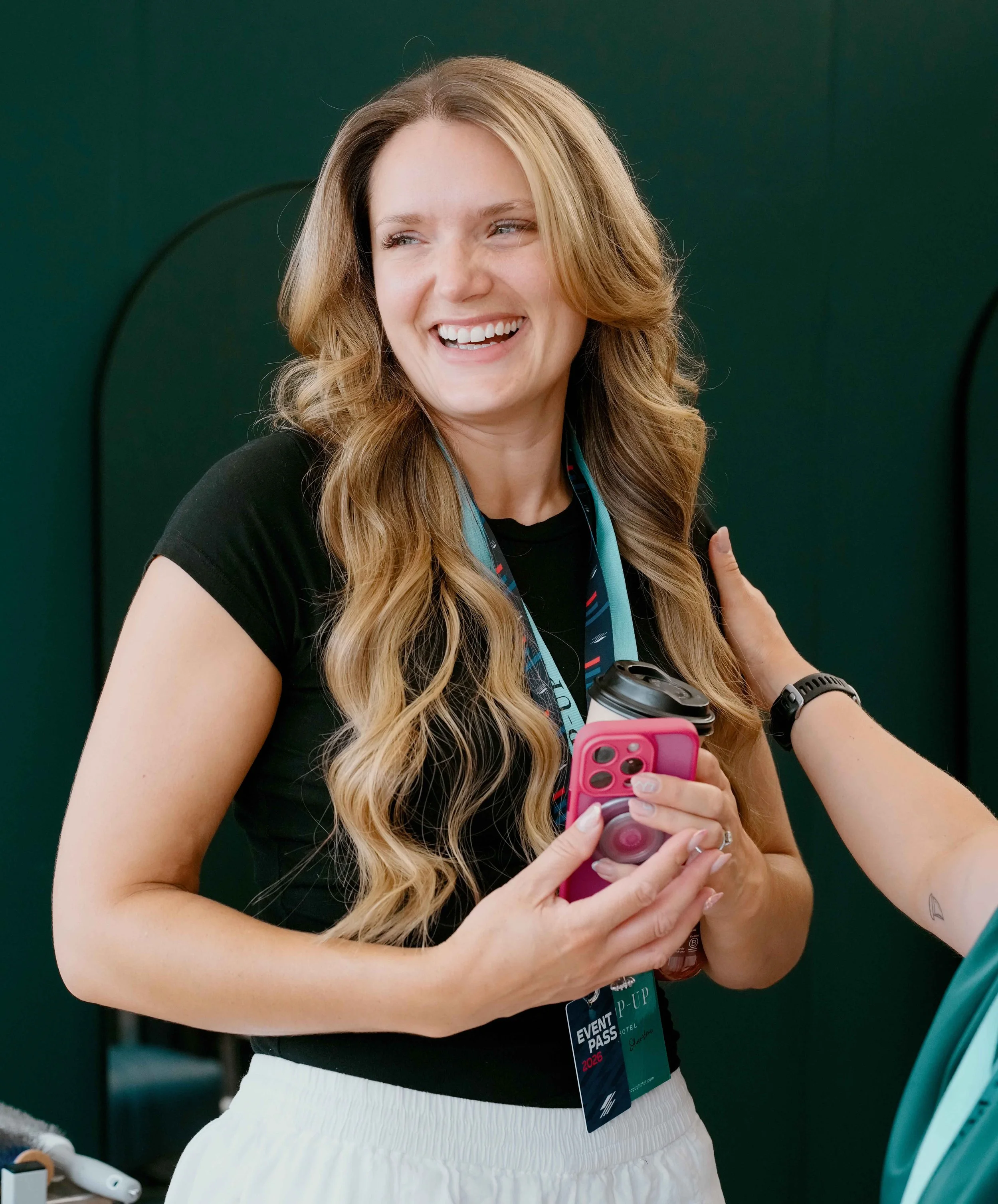 A smiling woman with long, wavy blonde hair holding a pink phone and a coffee cup, wearing a black shirt and a conference badge, standing against a dark green background.