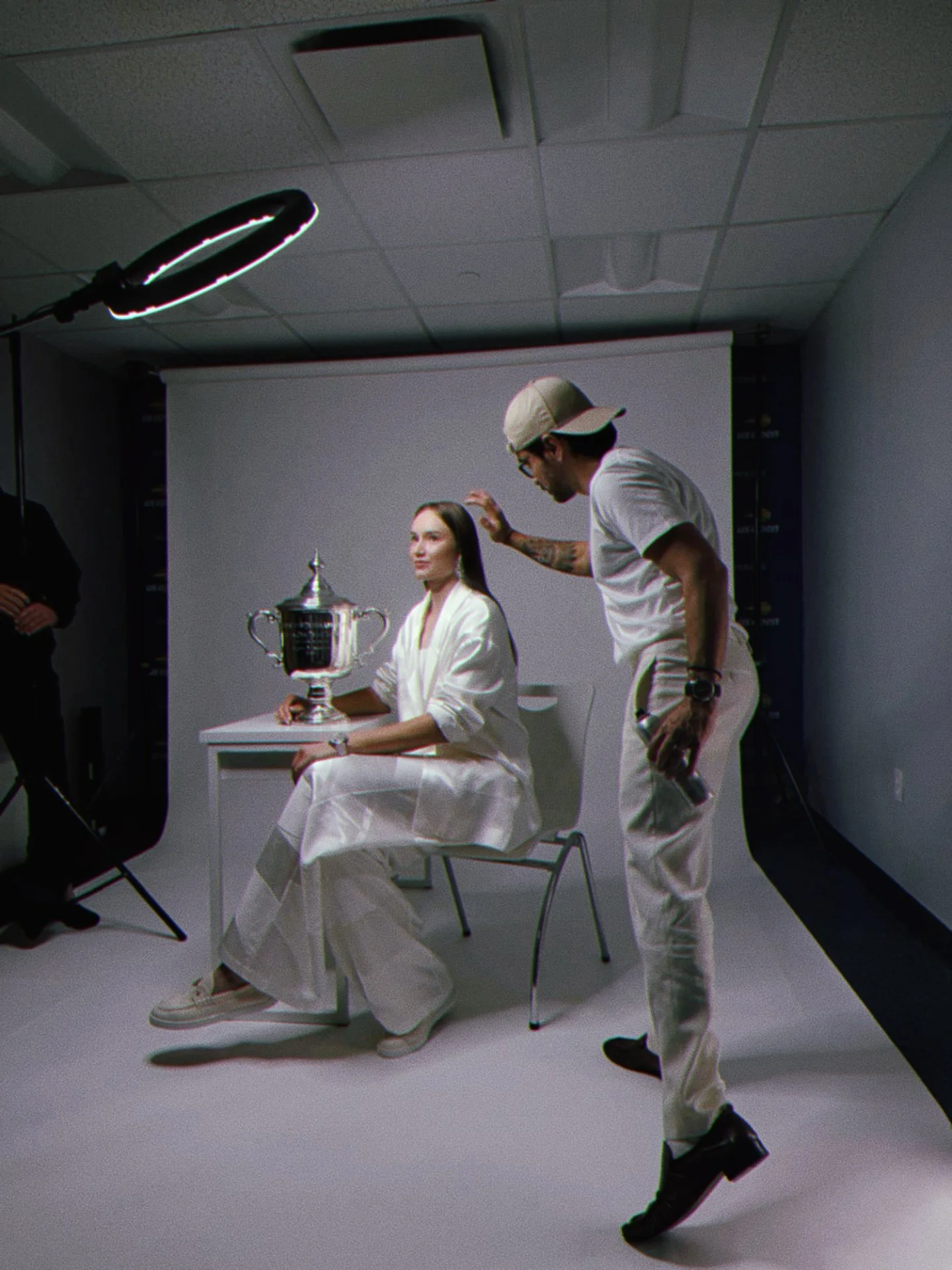 Elena Rybakina sitting in white clothing holding her trophy, with Marcos Verissimo standing beside her on the photoshoot a studio setting.